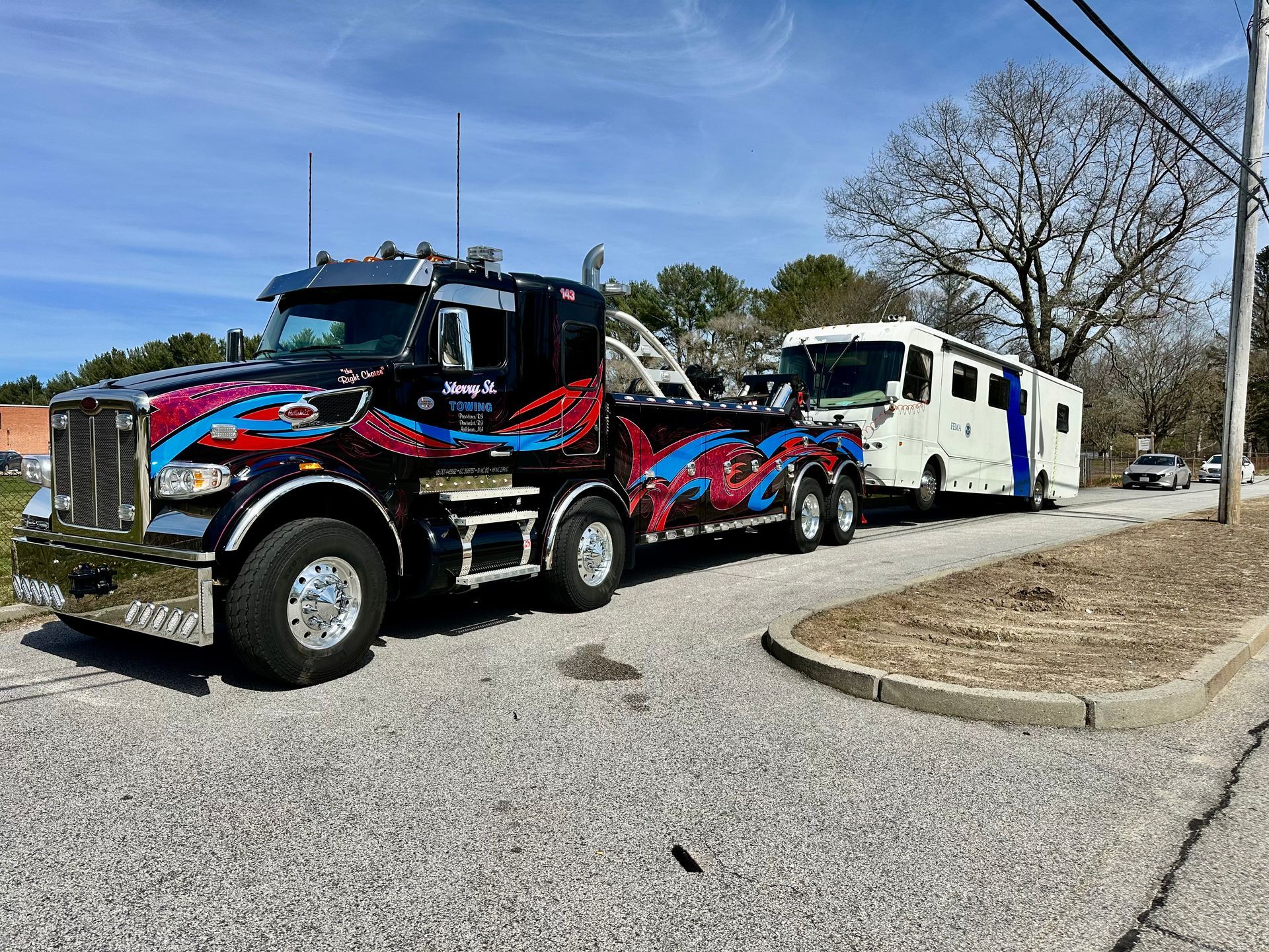 Black and red tow truck hauling a white semi-truck on a roadside under a clear blue sky.