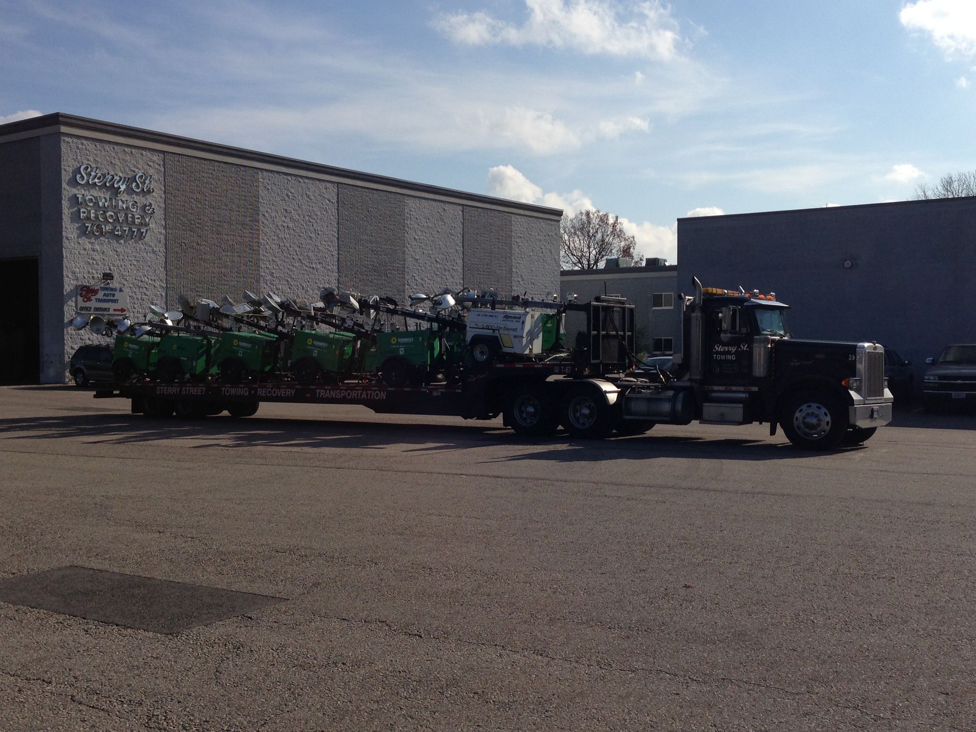 Black semi-truck hauling a flatbed trailer with green farm equipment in a parking lot.