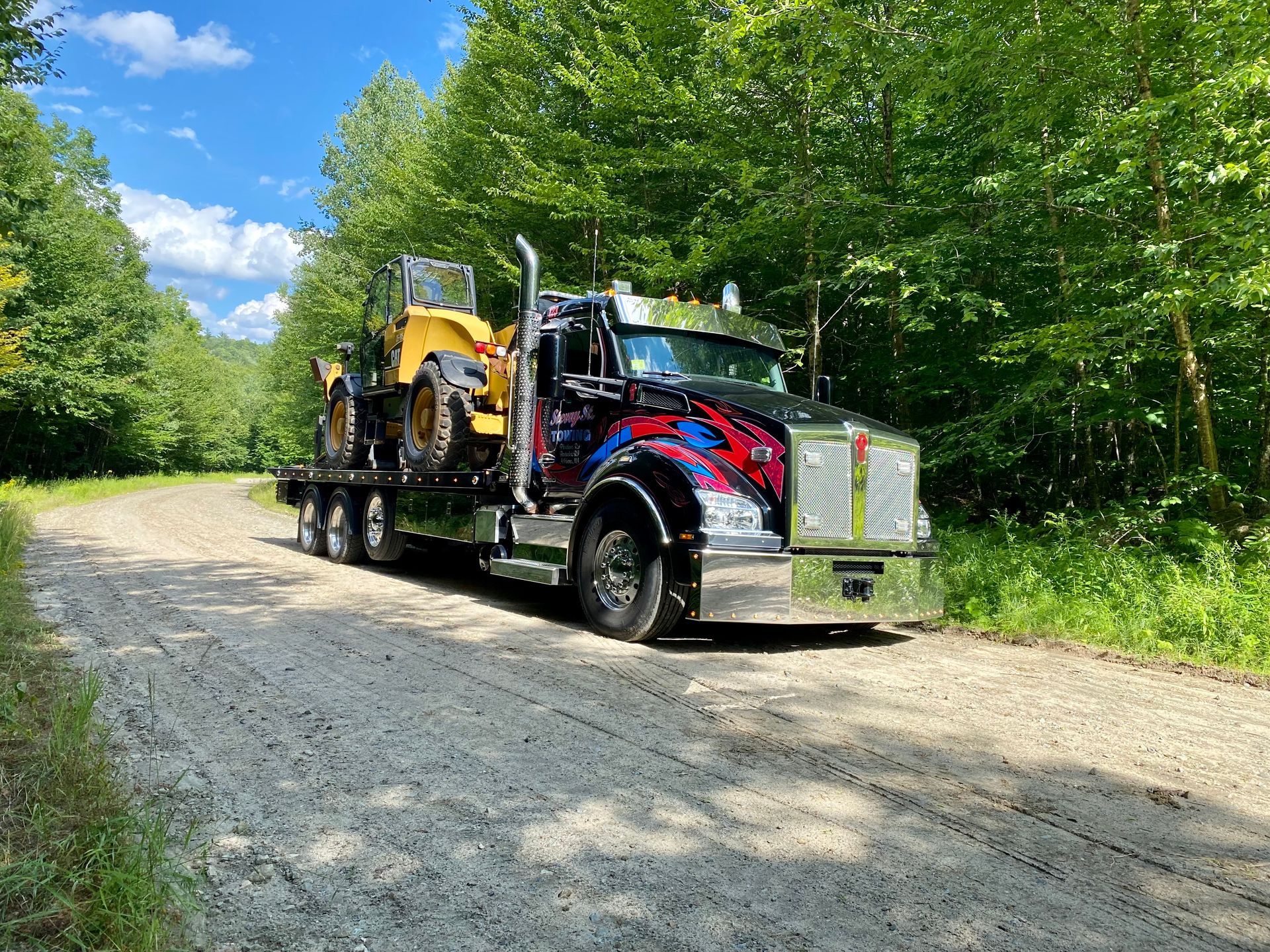 Red and chrome flatbed tow truck on a gravel road beside green trees under a blue sky.