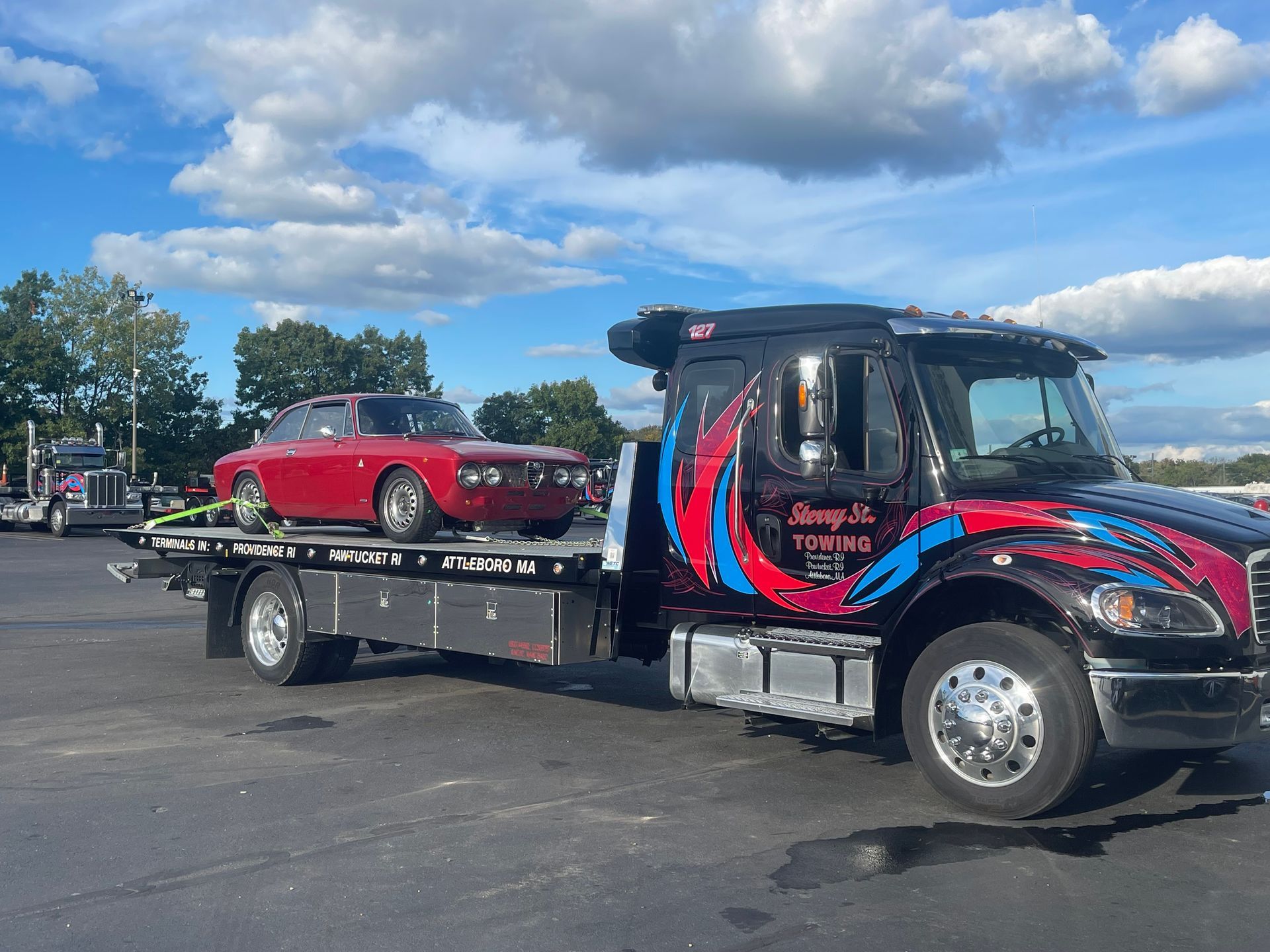 Black tow truck carrying a red classic car in a parking lot under a cloudy blue sky.