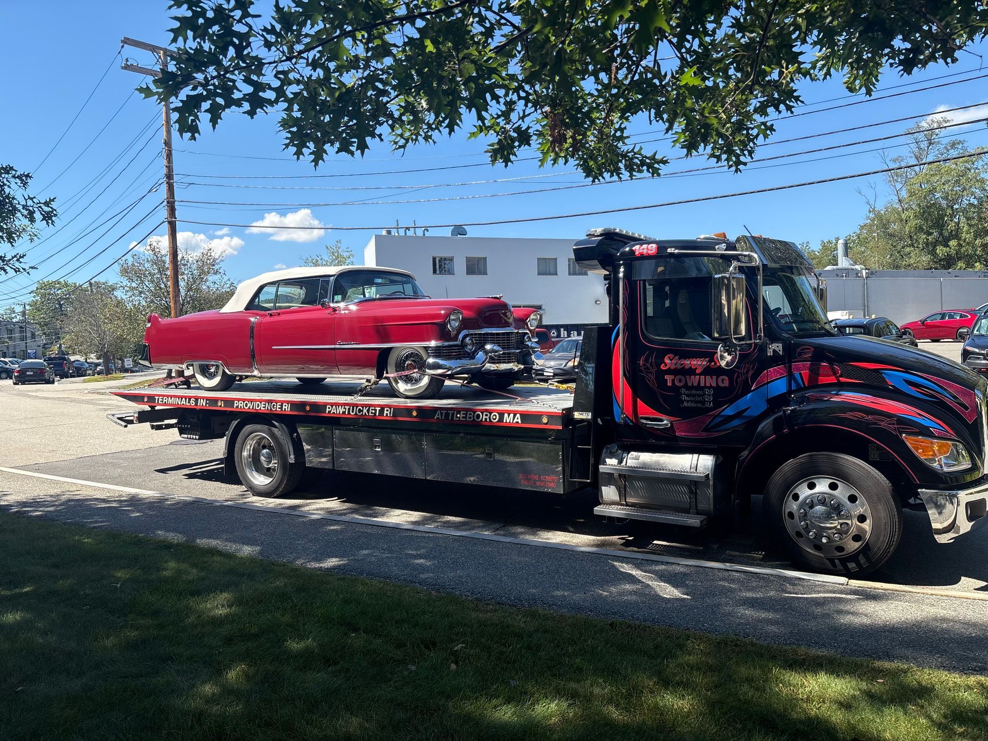 Tow truck hauling a red classic car on a flatbed along a sunny street.