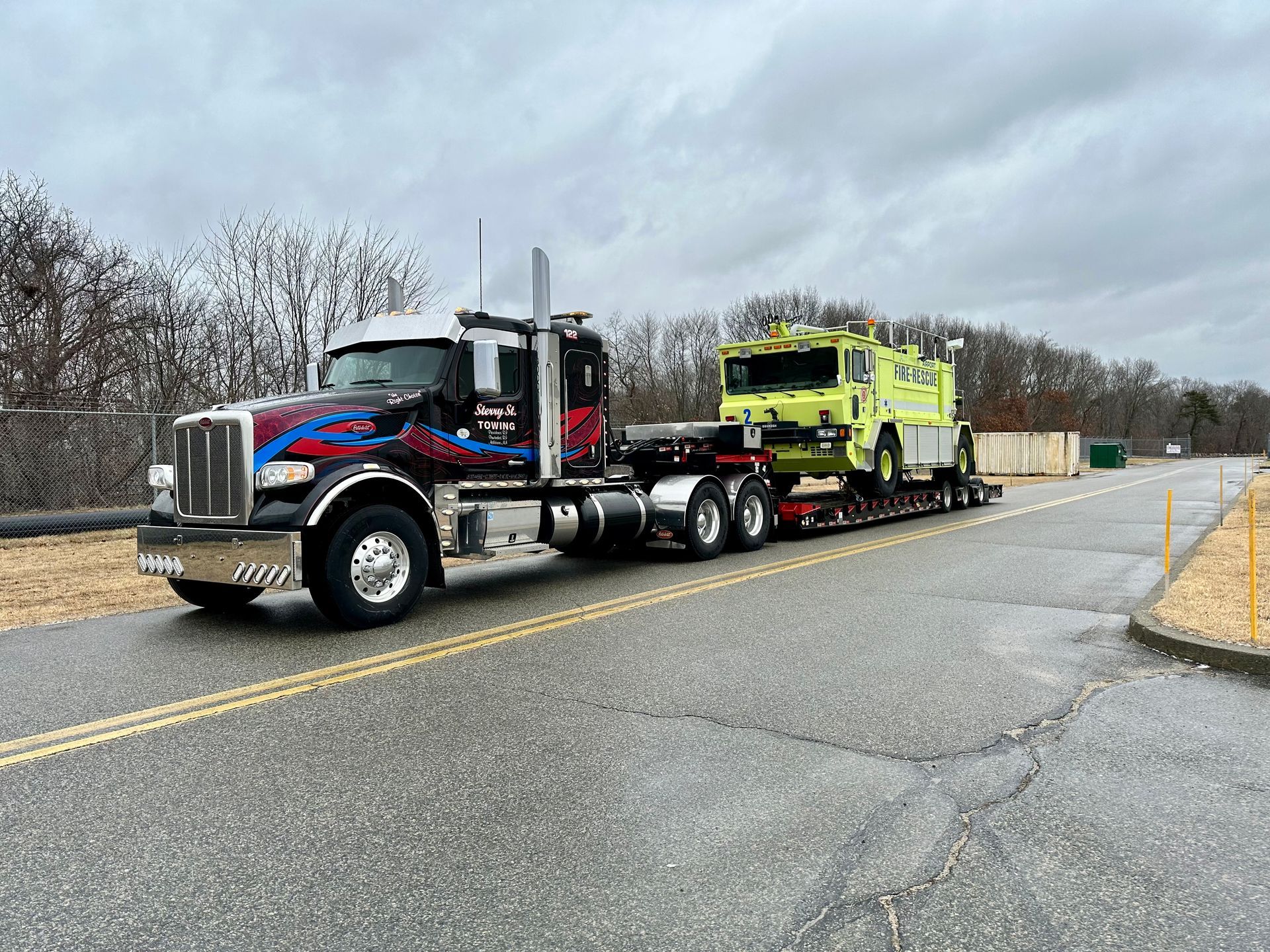Black semi truck towing a bright yellow fire truck on a road under cloudy skies.