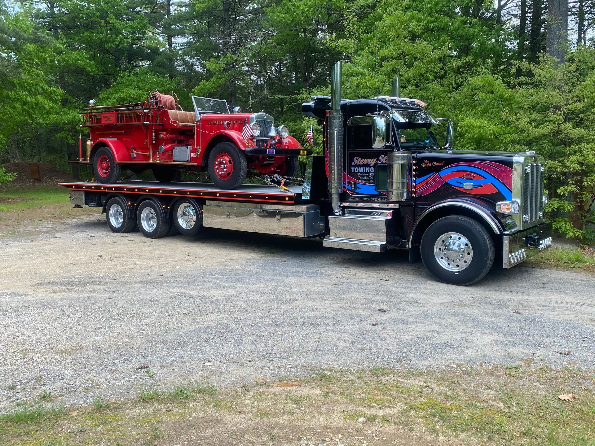 Red vintage fire truck on a flatbed tow truck in a gravel lot by trees.
