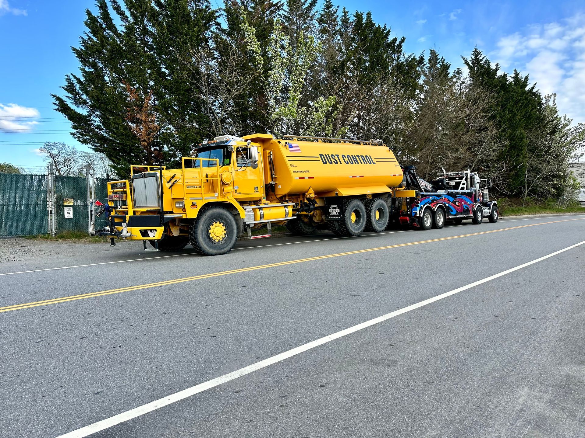 Yellow tanker truck parked on roadside with a blue utility vehicle and trees in the background.