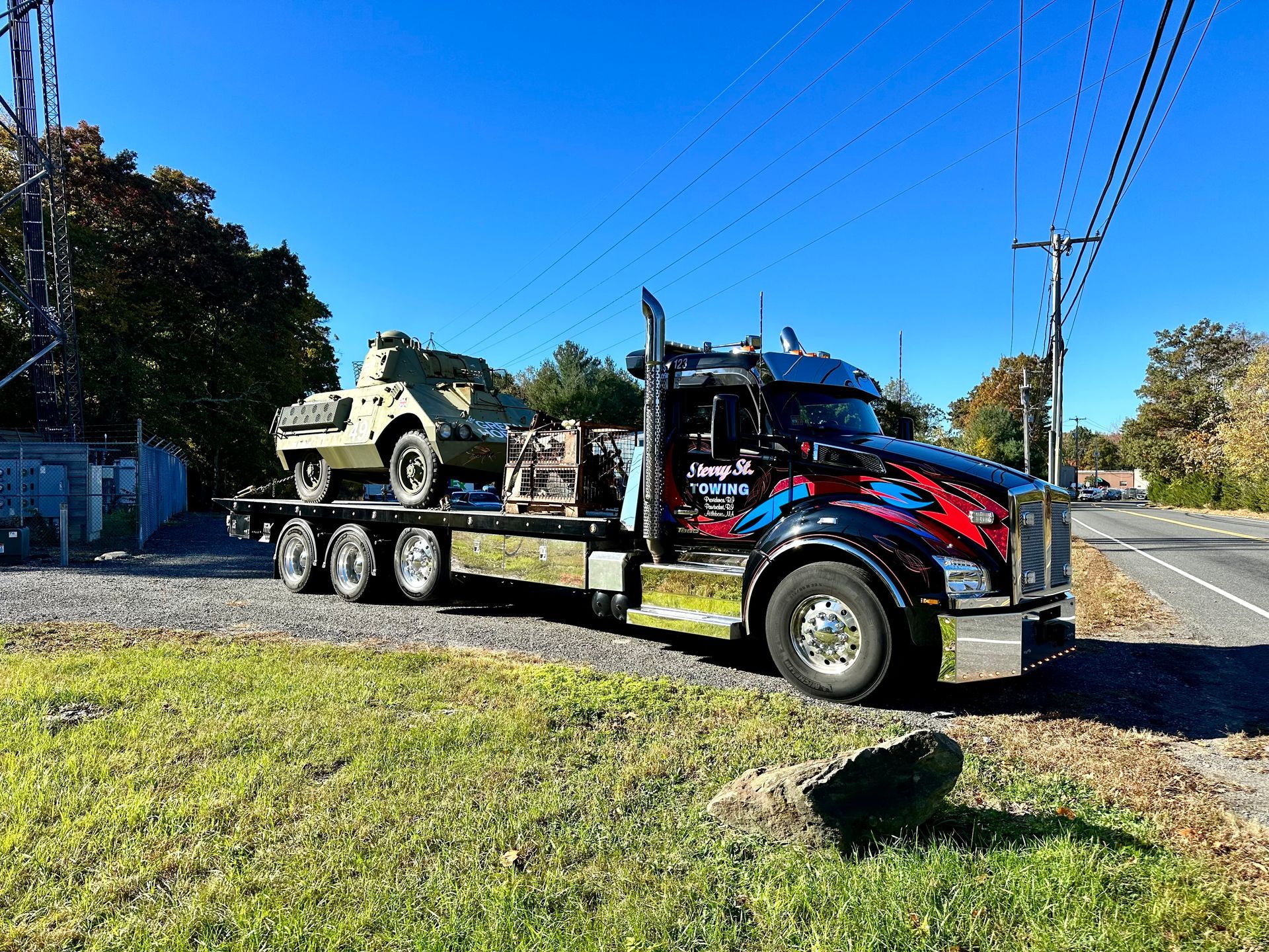 Semi-truck hauling a military-style vehicle on a flatbed beside a roadside under a clear blue sky.