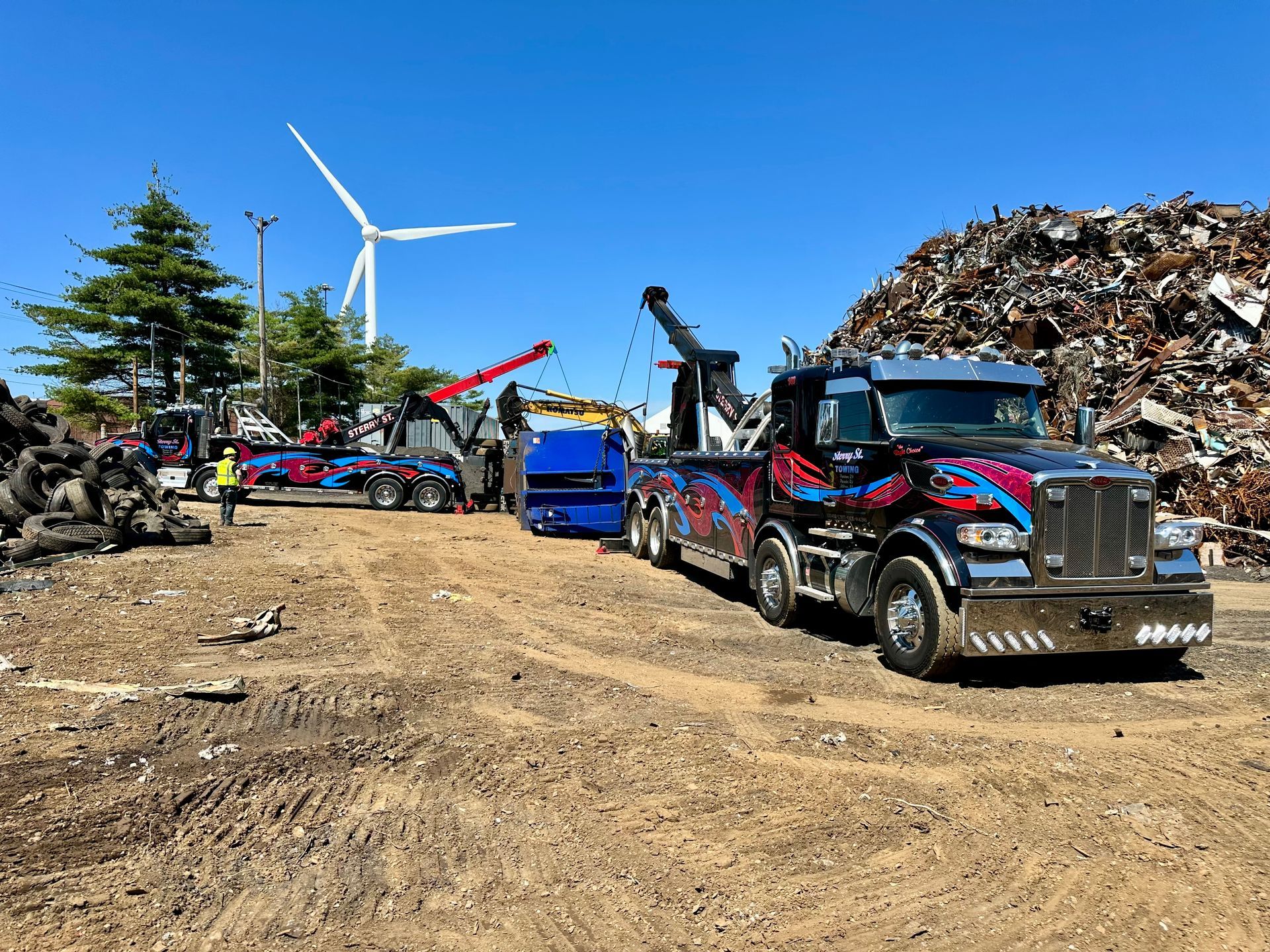 Tow trucks and cars lined up on a dirt lot under a clear blue sky with a wind turbine in the background.