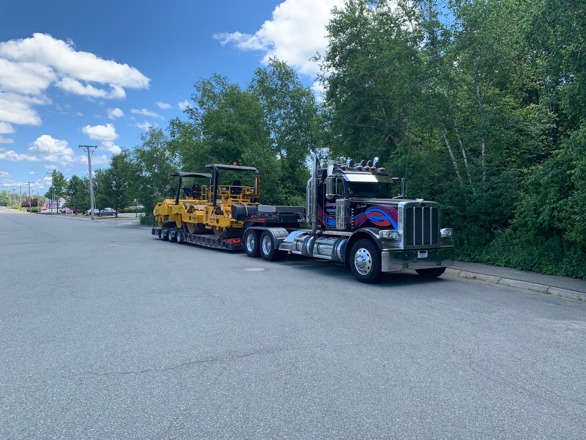 Black semi-truck hauling yellow road equipment on a gravel lot beside trees and a cloudy blue sky.