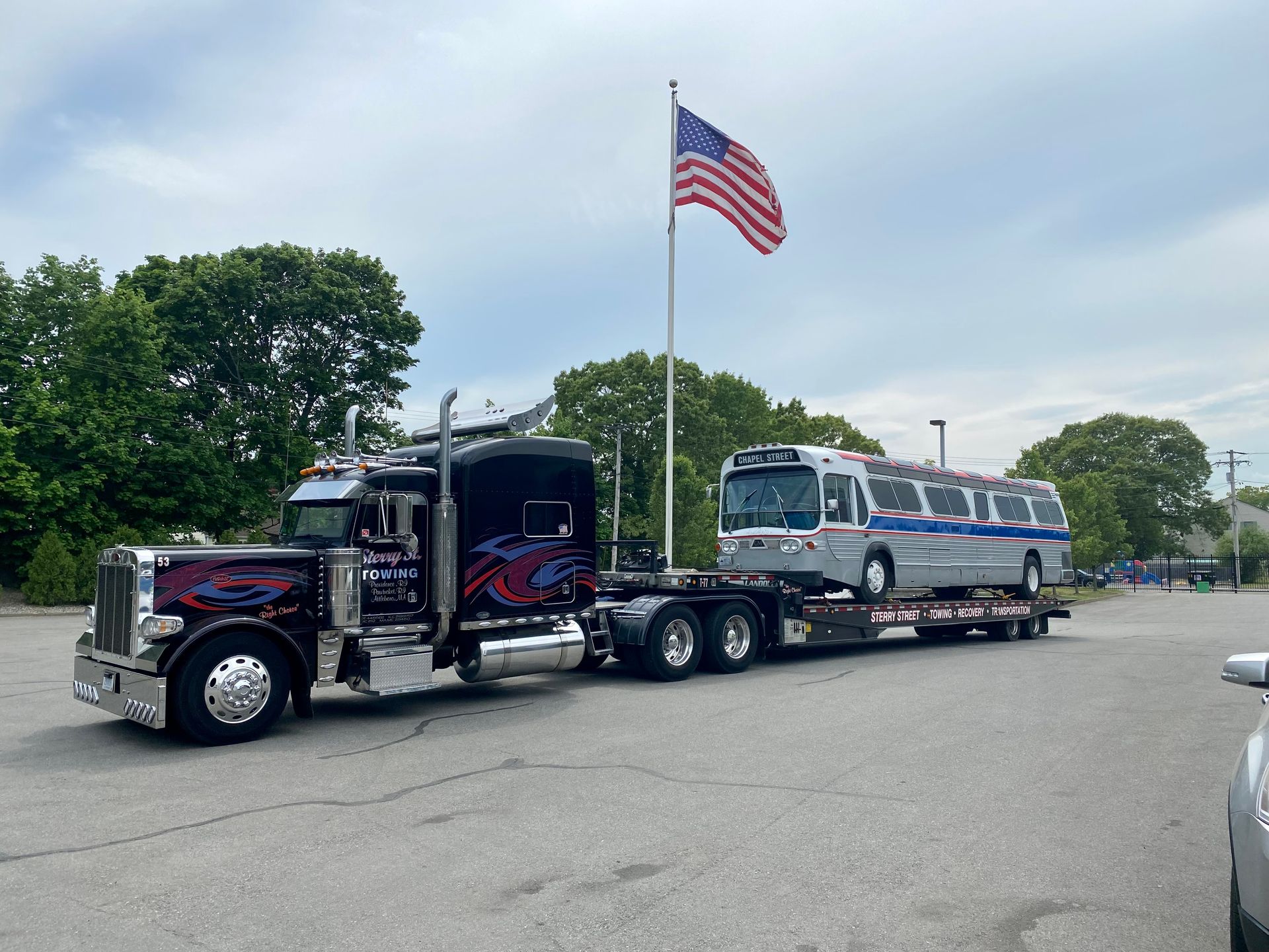 Black semi truck towing a blue bus on a road, with a large U.S. flag waving above it.