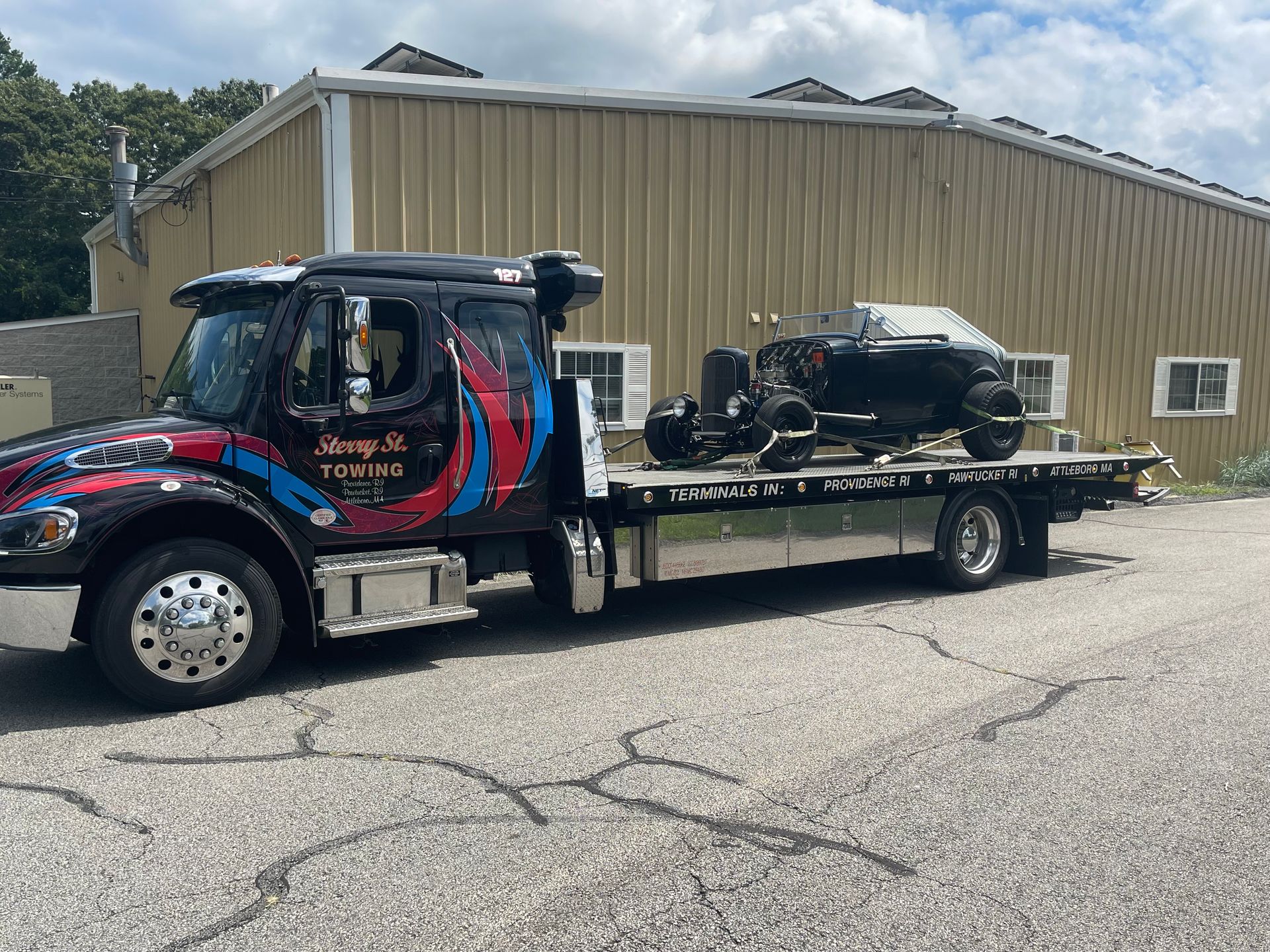Tow truck hauling a black car beside a beige industrial building on a gravel lot.