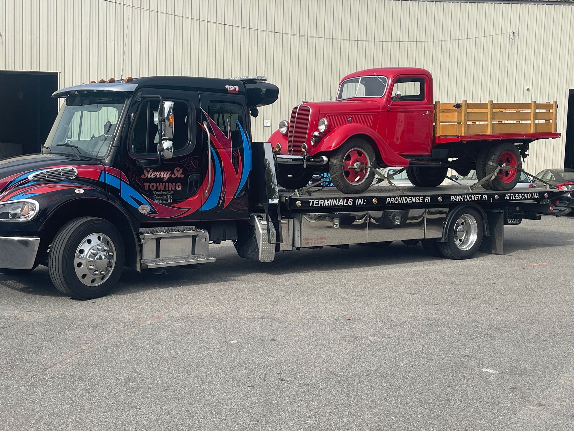 Red vintage truck on a flatbed tow truck, parked outdoors on a street.