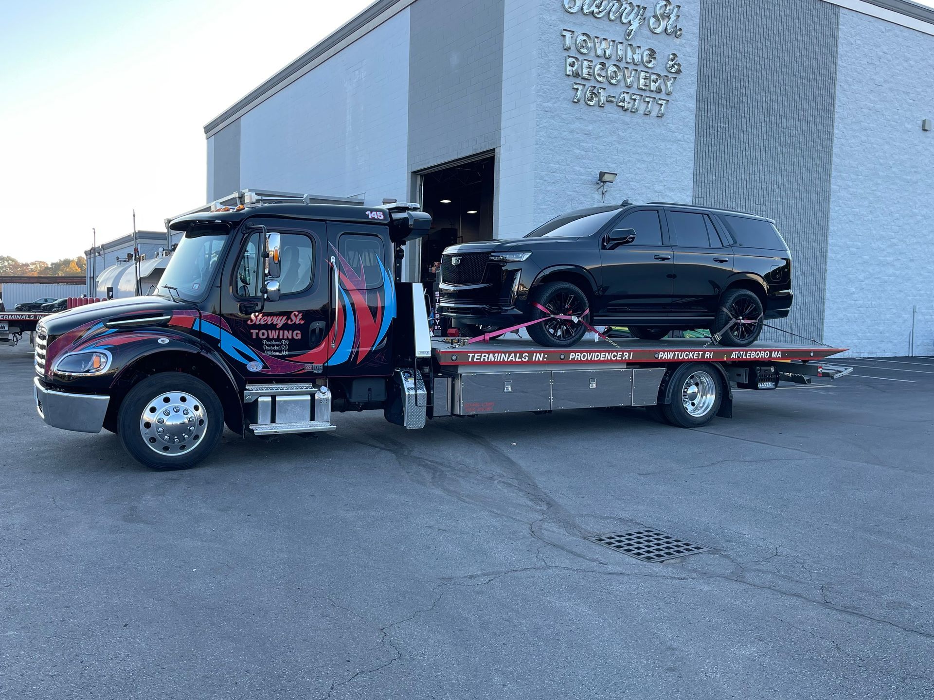 Tow truck hauling a black SUV beside a warehouse on a paved lot.