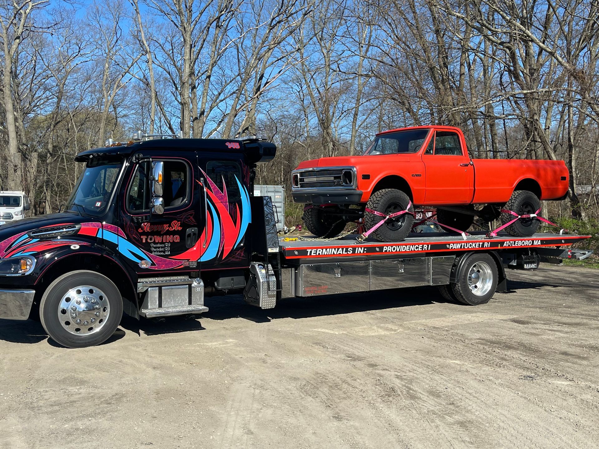 Tow truck hauling a bright orange pickup on a flatbed in a dirt lot.