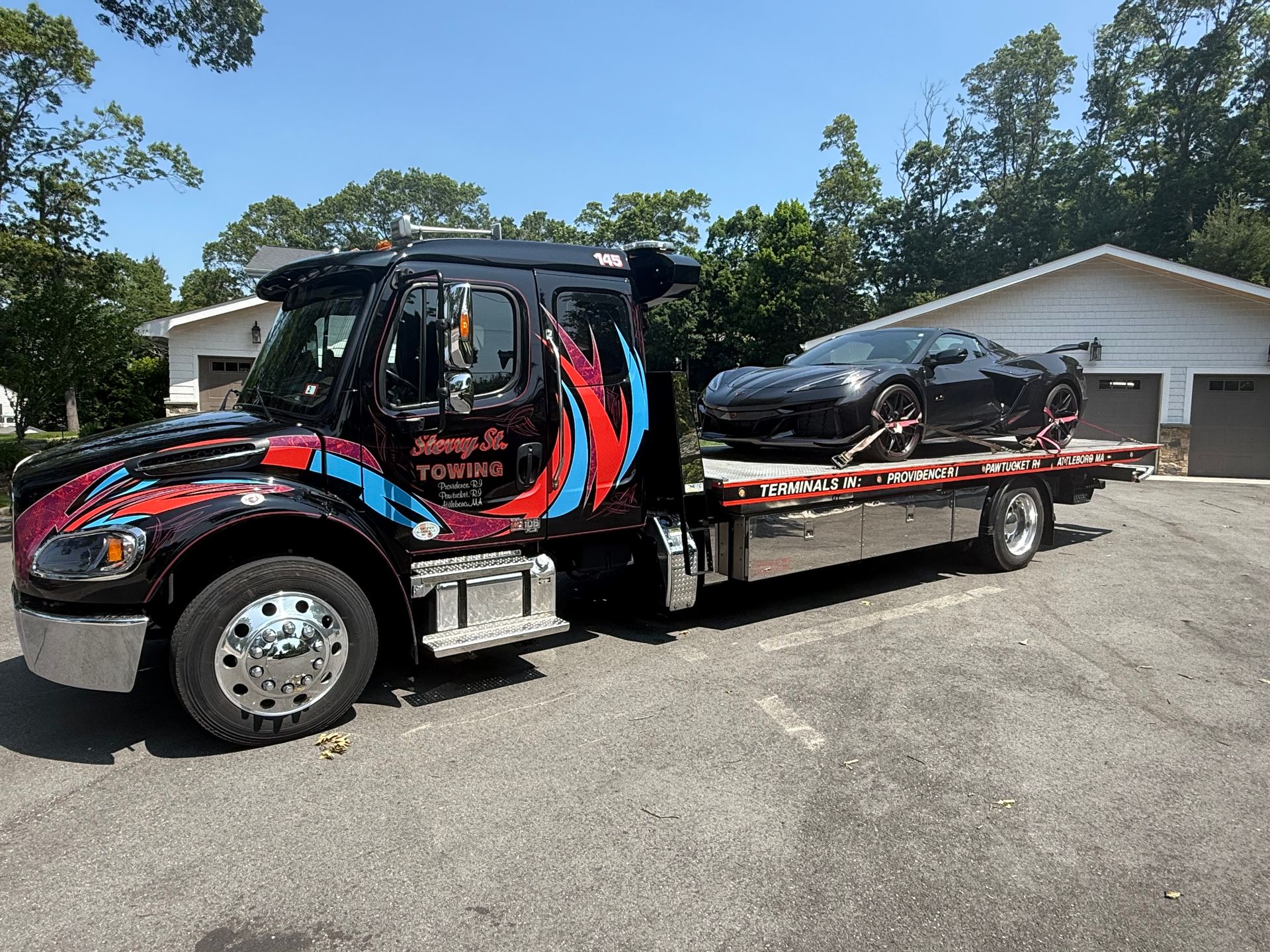 Tow truck hauling a black car on a flatbed in a gravel lot, with houses and trees behind.