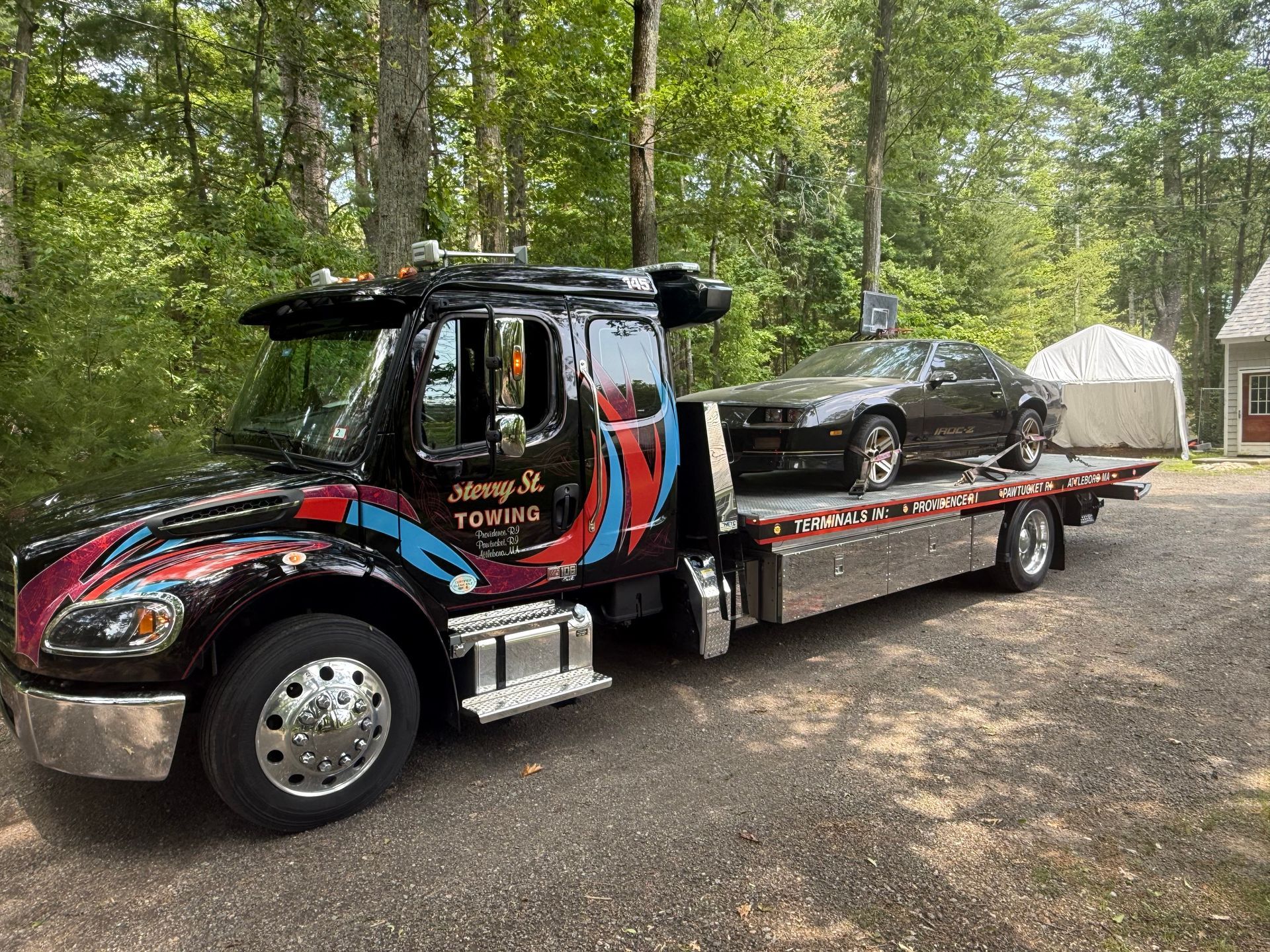 Tow truck hauling a black car on a flatbed in a wooded campsite area.