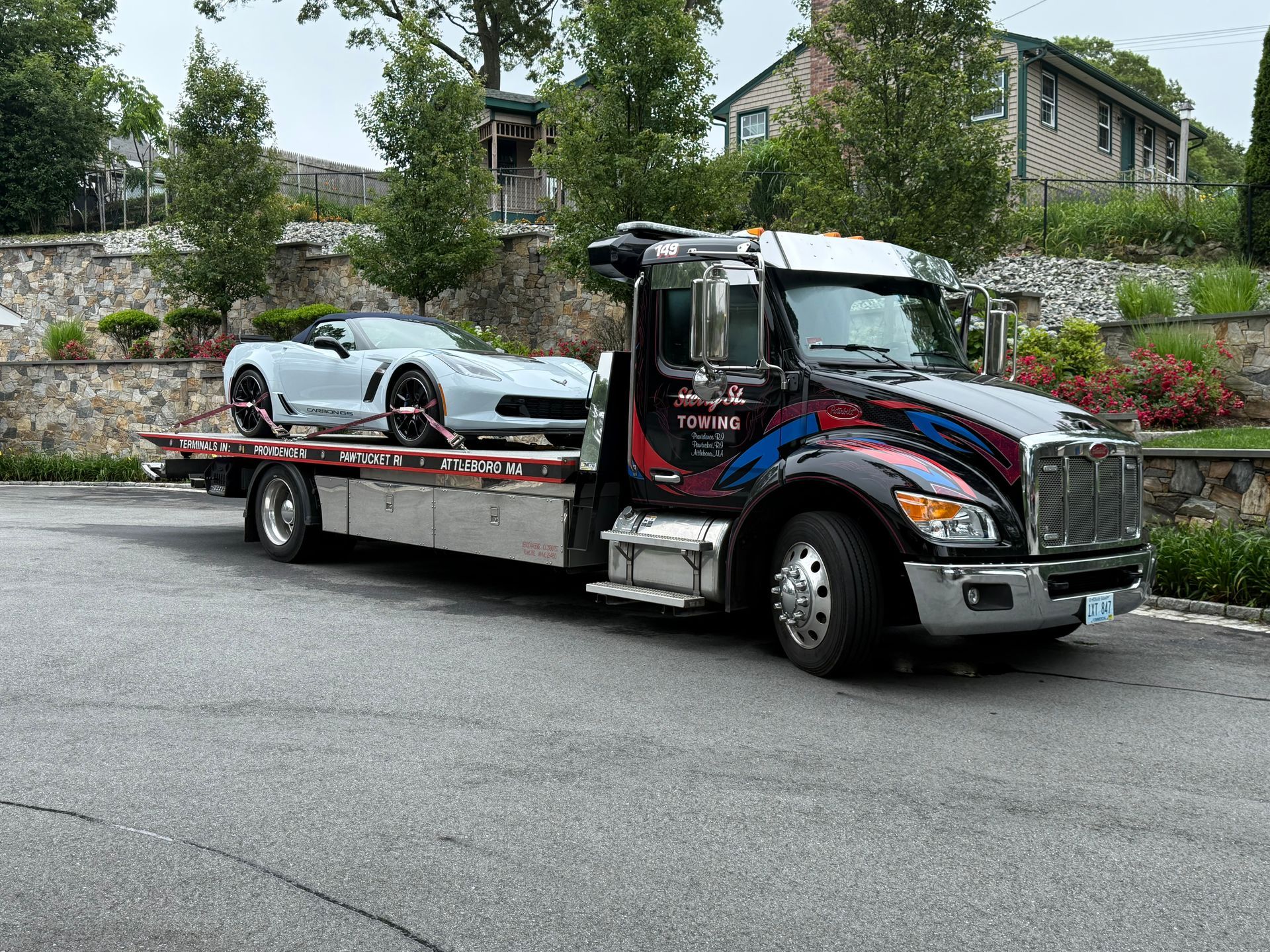 Tow truck carrying a white sports car on a flatbed in a residential area.