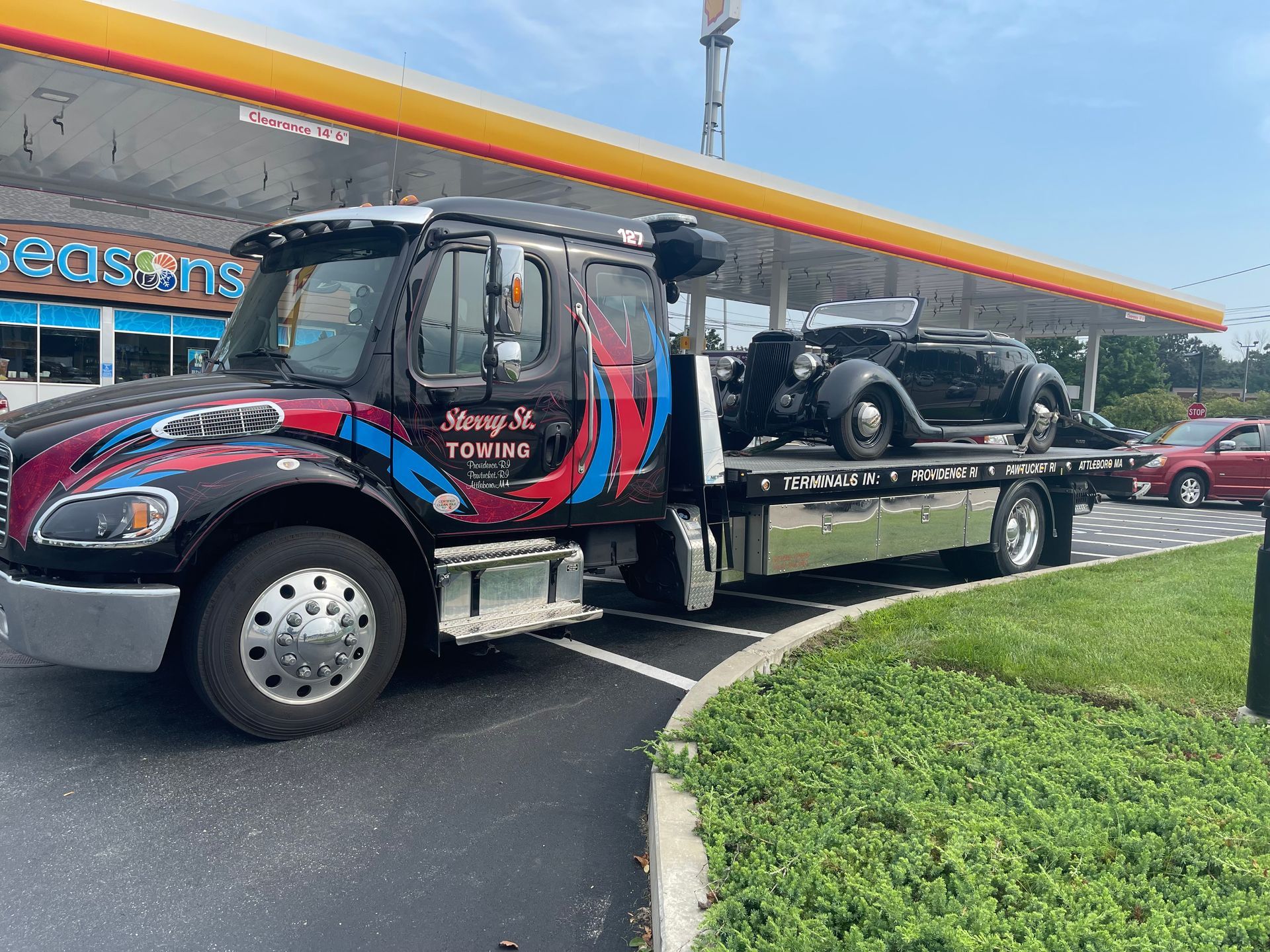 Tow truck carrying a vintage black car at a gas station, with red and blue truck cab and green grass nearby.