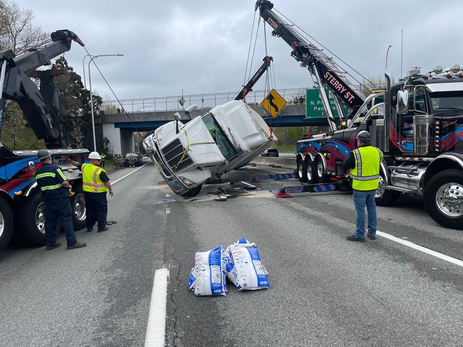 Overturned truck on highway after crash, with tow trucks, workers, and water jugs spilled on the road.