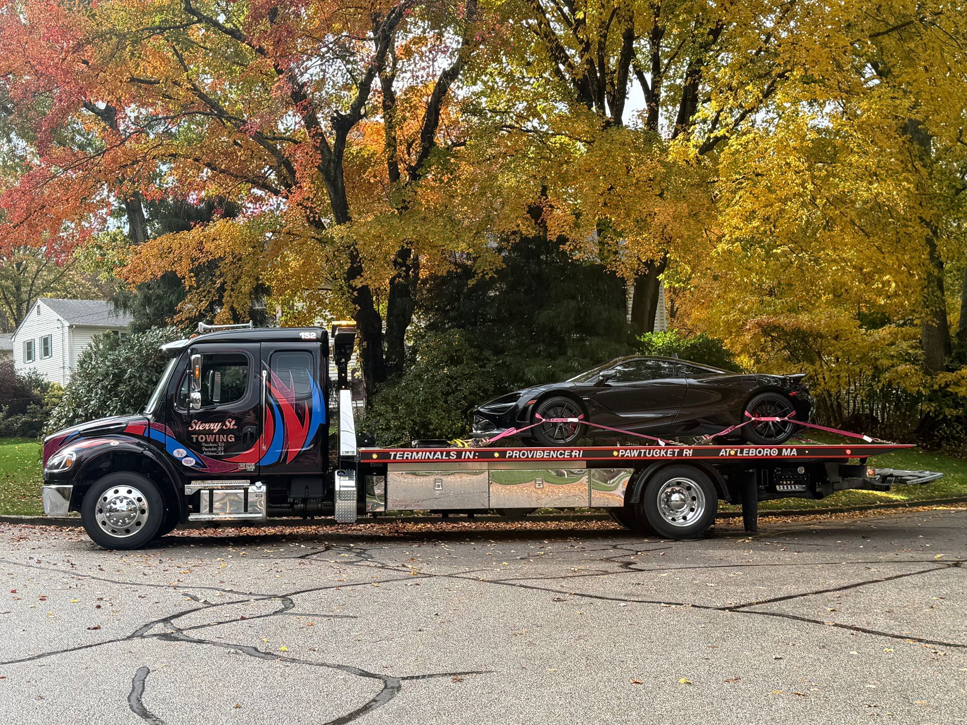 Tow truck carrying a black sports car on a flatbed, parked on a road with autumn trees behind.