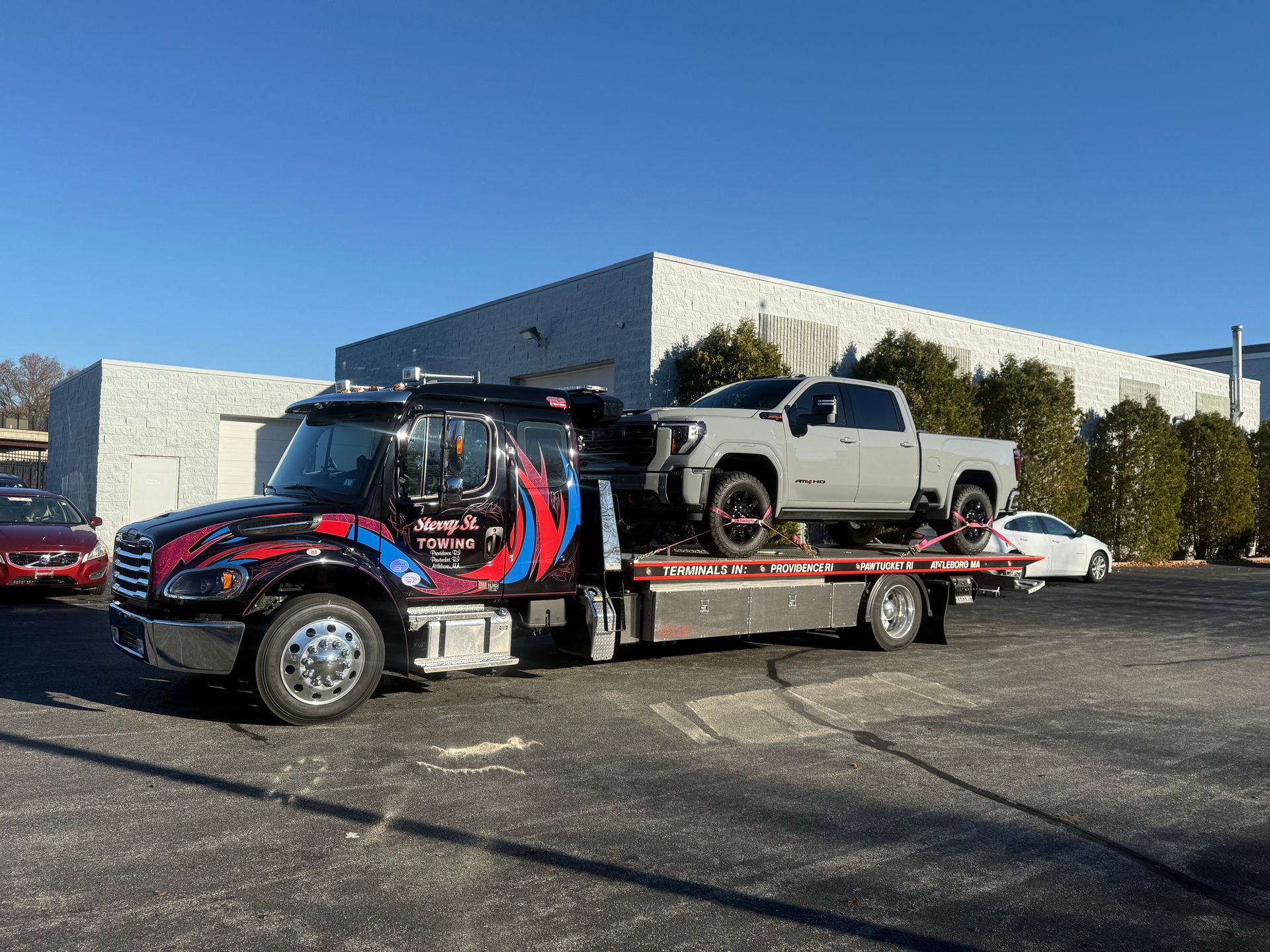 Flatbed tow truck carrying a silver pickup in a parking lot on a sunny day.