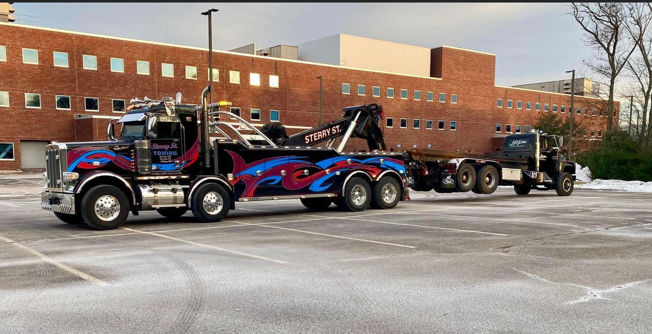 Tow truck hauling a loaded trailer in a snowy parking lot beside a brick building.