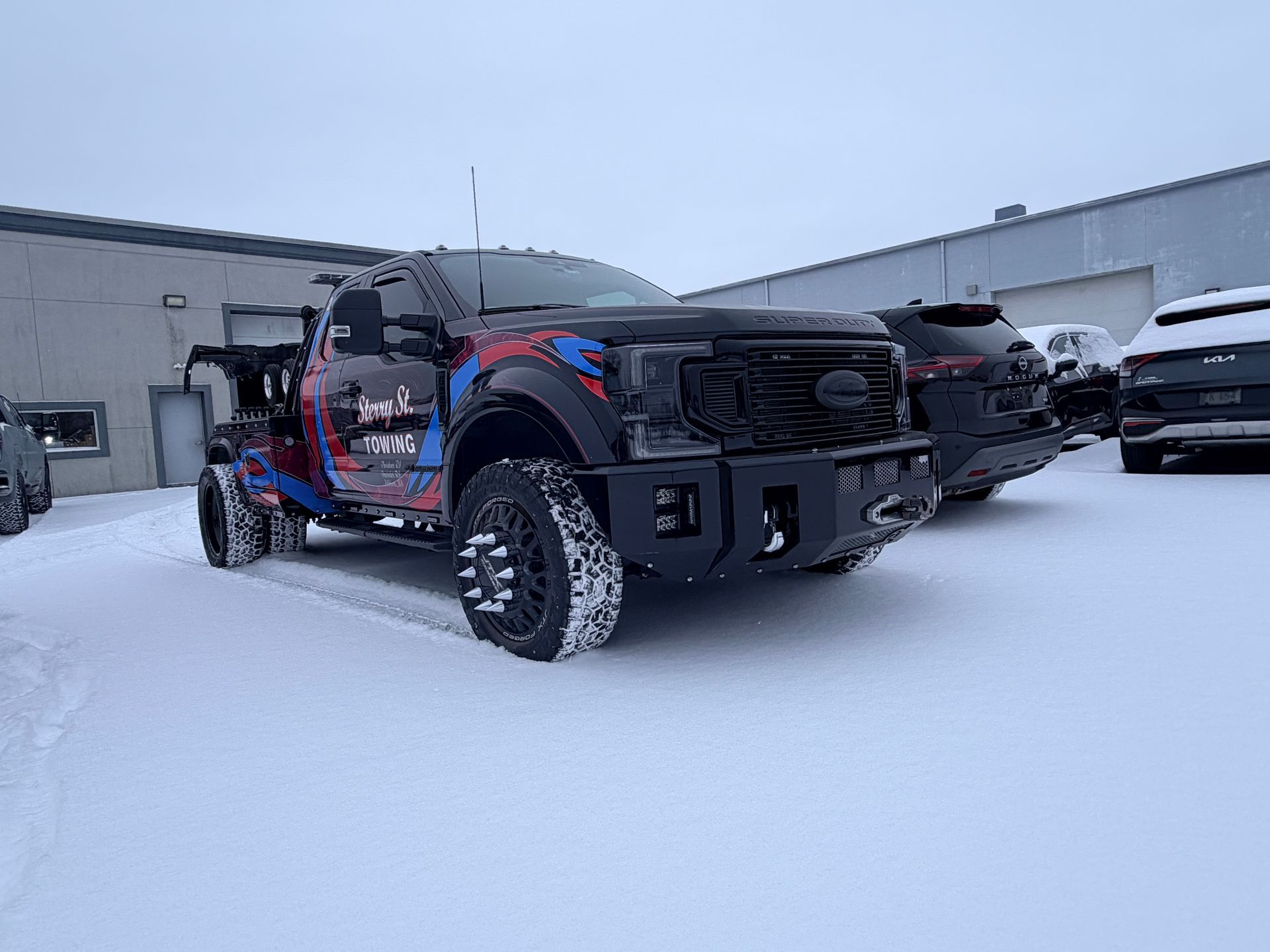 Black off-road truck with red accents parked in a snowy lot beside industrial buildings.