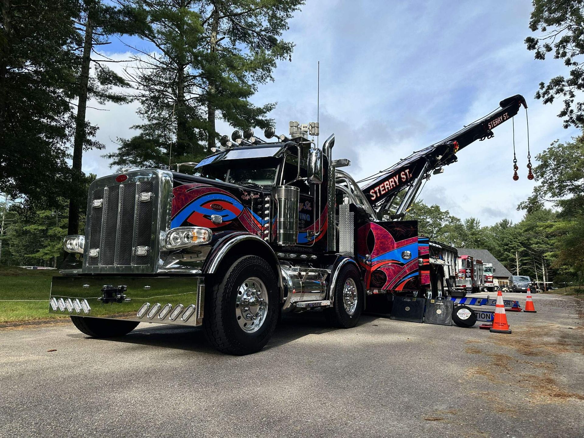 Custom chrome tow truck with raised boom parked on gravel, with cones and trees nearby