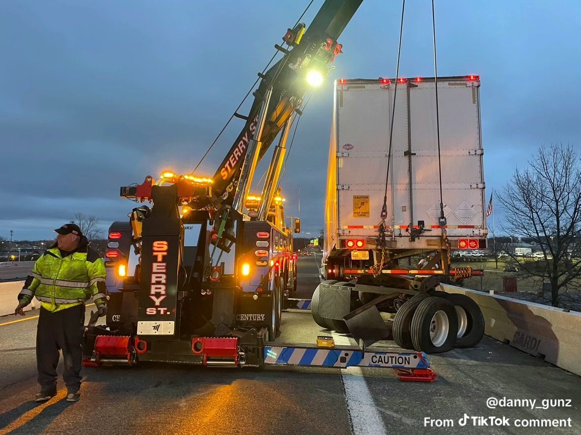 Tow truck lifting a white semi-trailer on a wet highway at dusk, with a worker nearby.