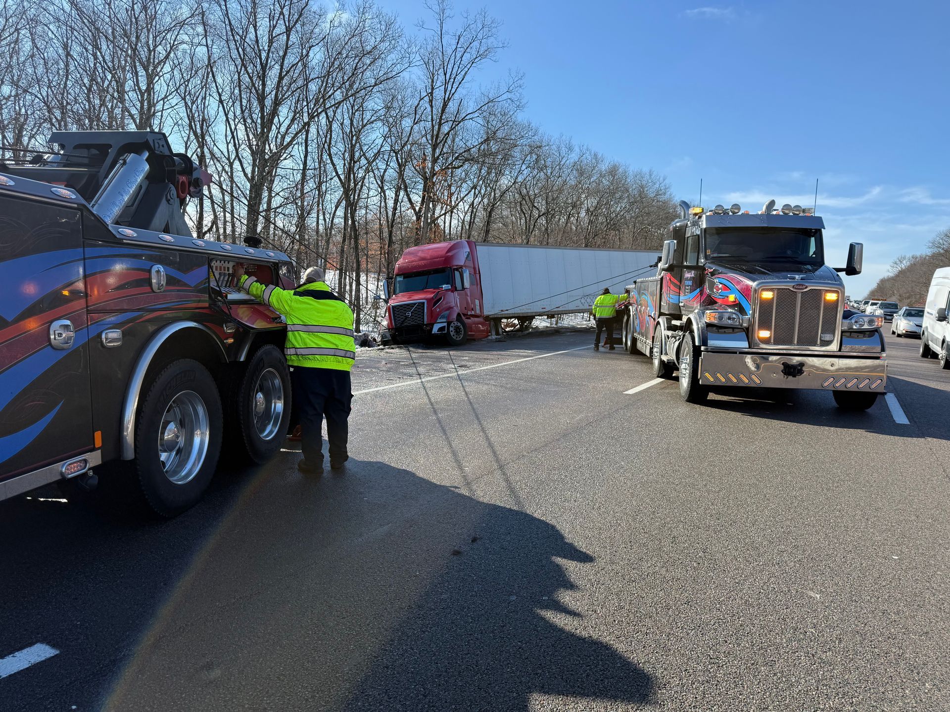 Tow trucks and a worker blocking a highway near a red semi-truck and trailer under a clear sky.