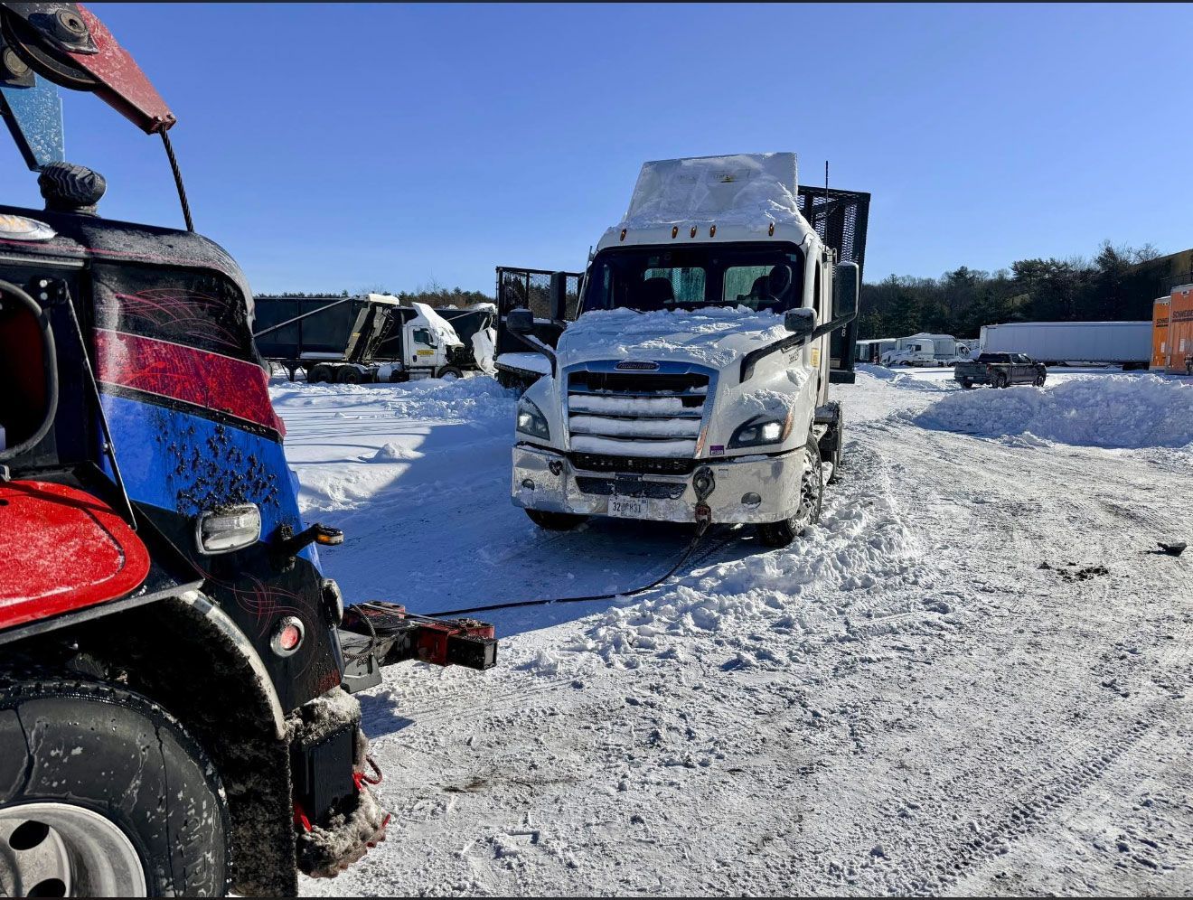 Snow-covered lot with a white semi-truck centered and a red vehicle at left under a clear blue sky.
