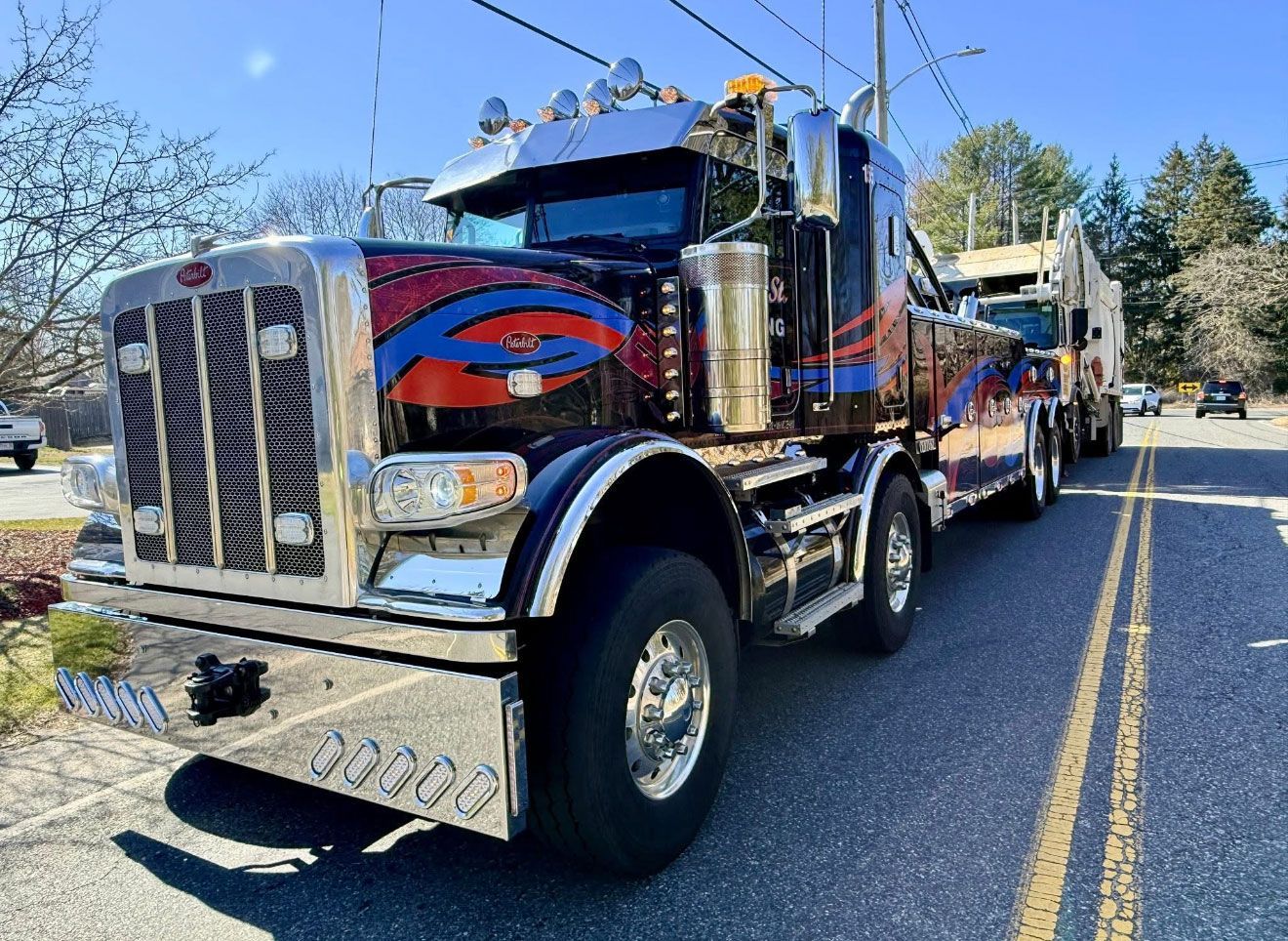 Blue semi-truck with chrome details parked on a road with double yellow lines and trees in the background.