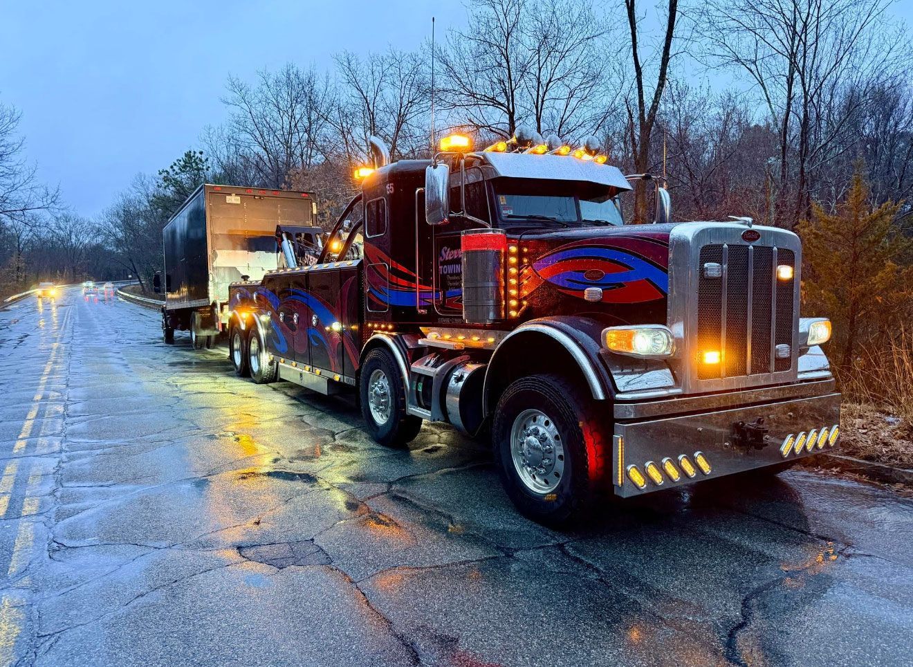 Red semi-truck hauling a trailer on a wet, snowy roadside at dusk, with headlights and amber lights on.