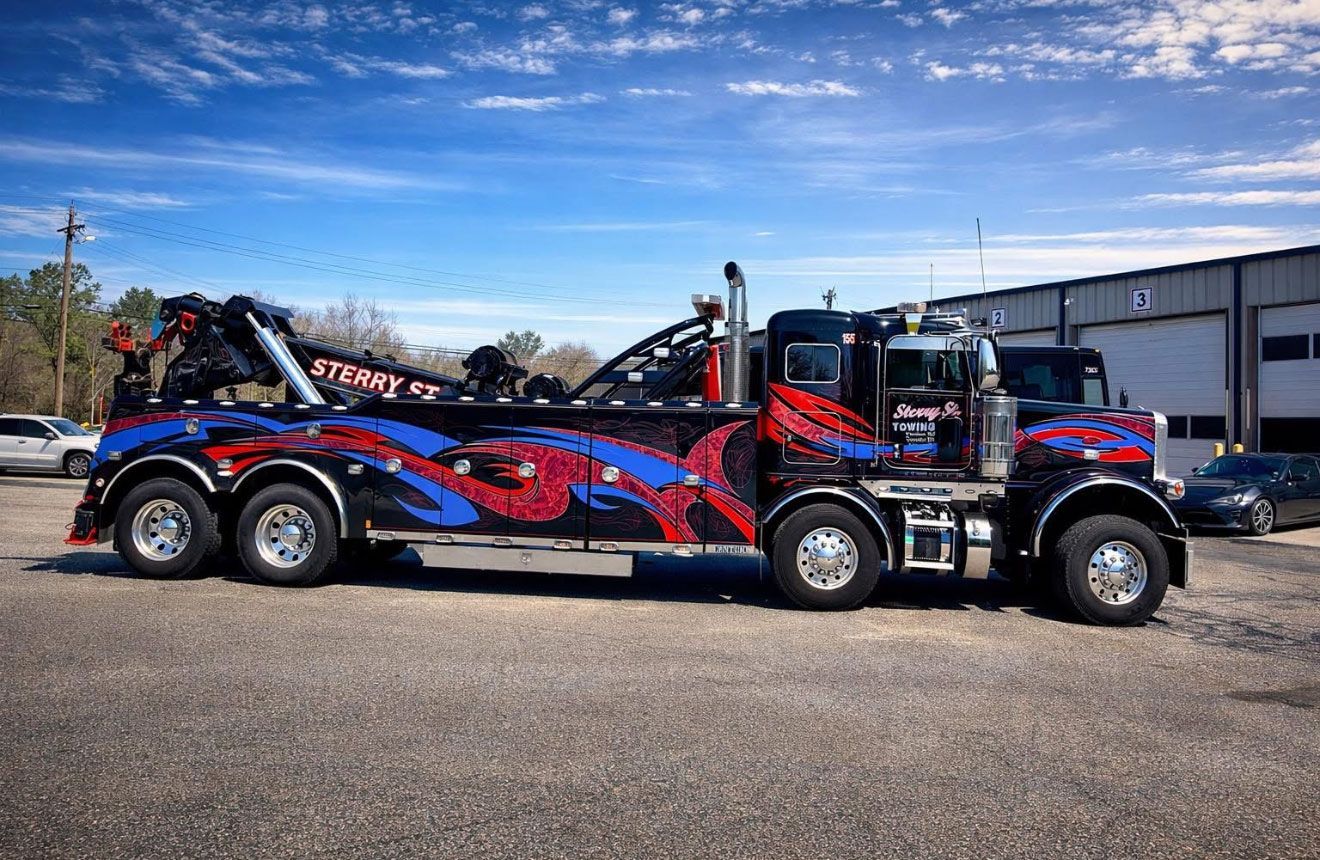 Red and black tow truck with blue flame graphics parked in a lot on a sunny day.