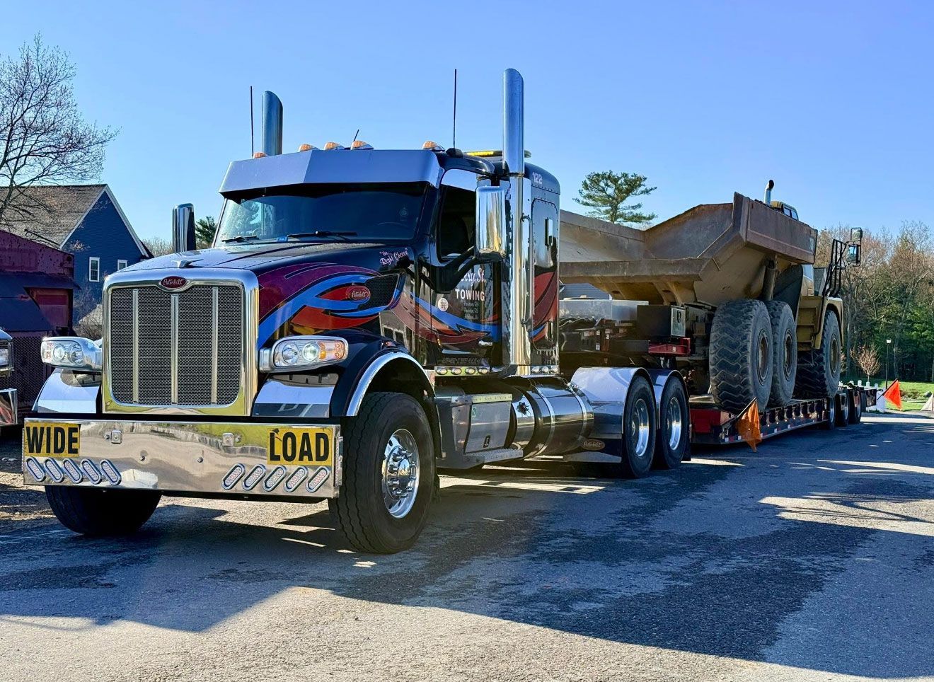 Red wide-load semi truck hauling a large house along a road, with escort cones and clear blue sky.