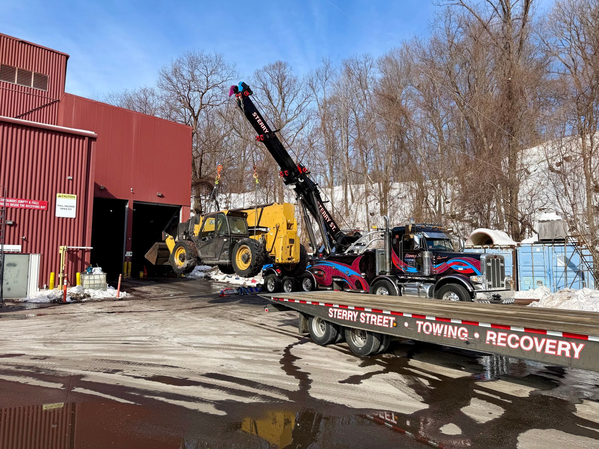 Tow truck with raised boom and car carrier in a snowy lot beside a red warehouse.