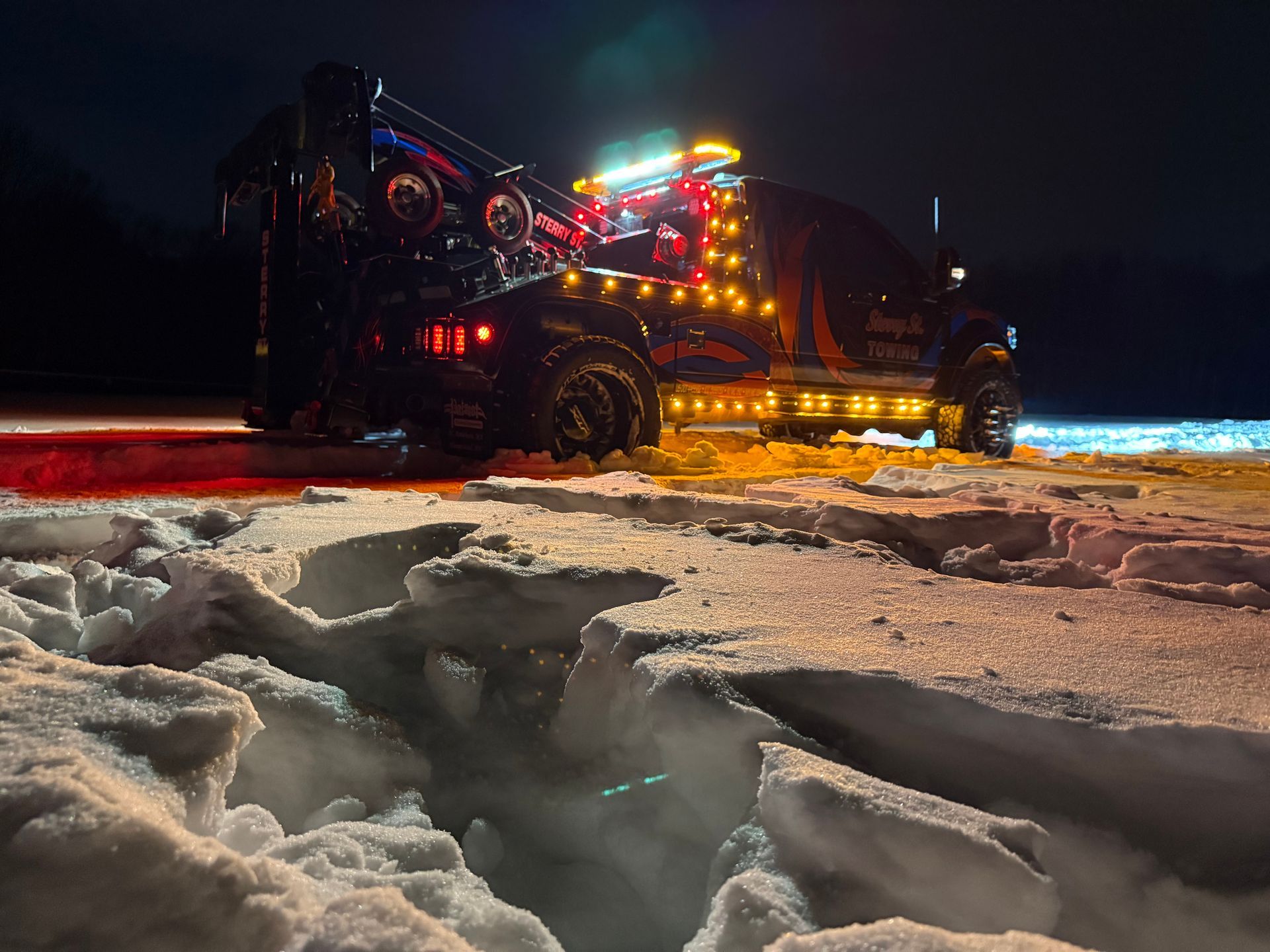 Tow truck with flashing lights pulling a car from icy snow at night.