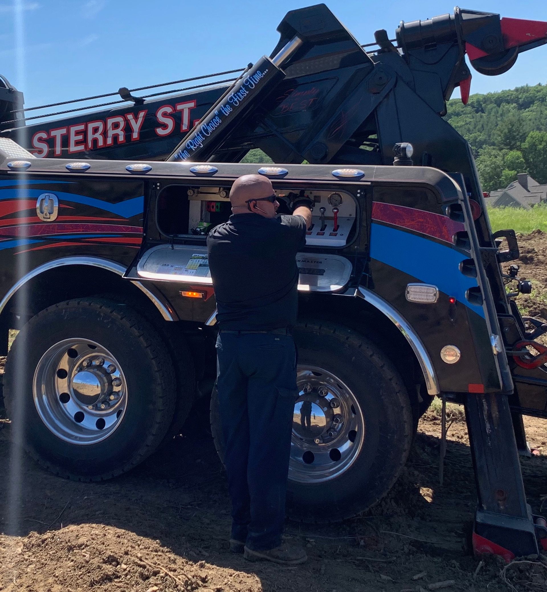 Worker standing beside a black tow truck with red and blue stripes, adjusting equipment outdoors.