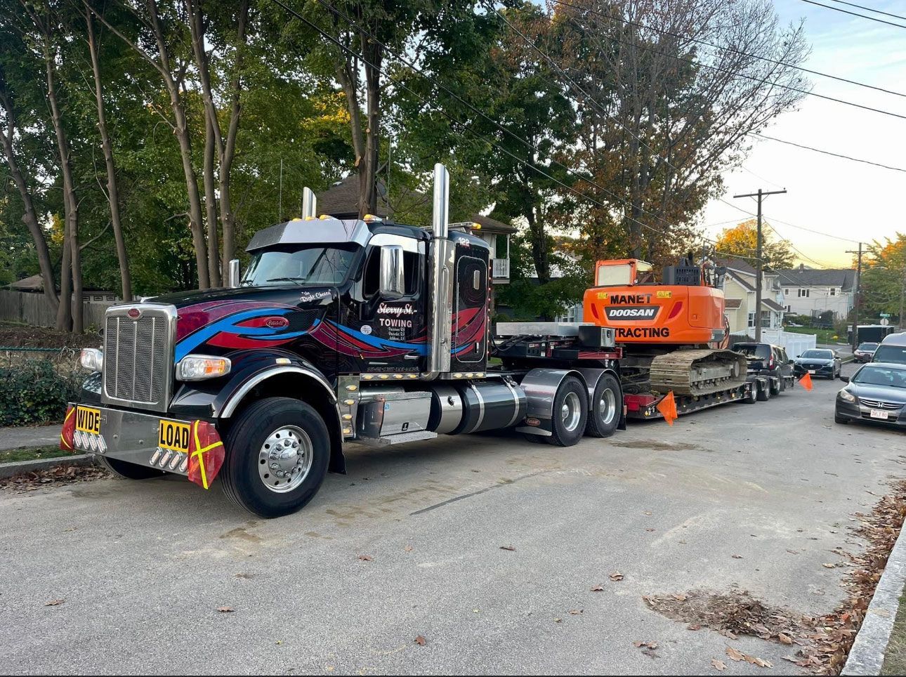 Black semi-truck hauling orange equipment on a residential street lined with trees and parked cars.