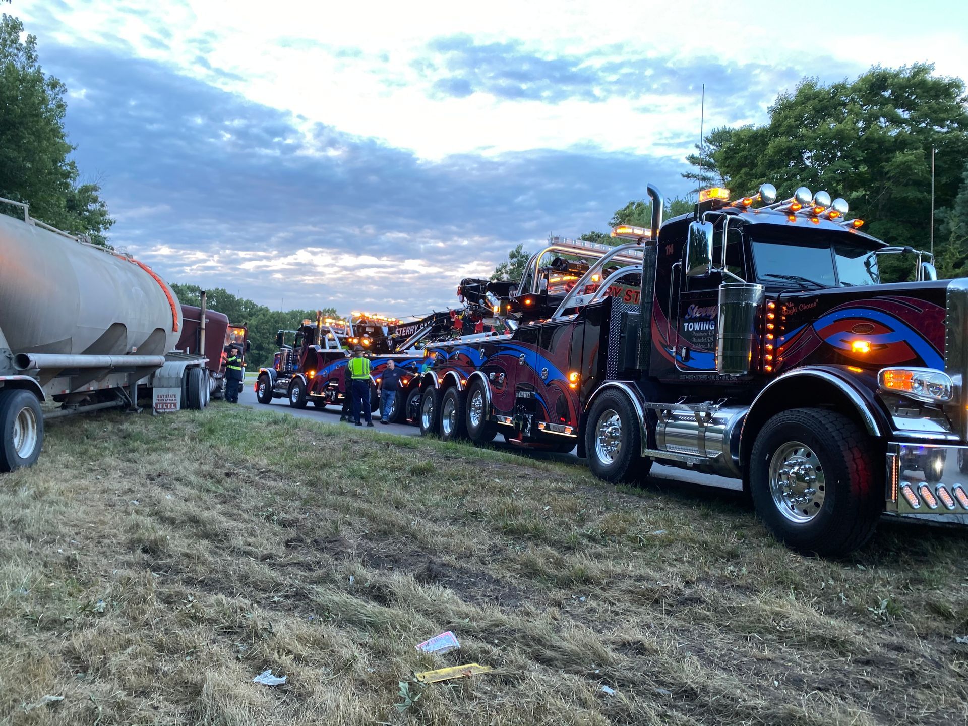 Line of decorated semi trucks at dusk on a grassy roadside, with a tanker truck on the left.