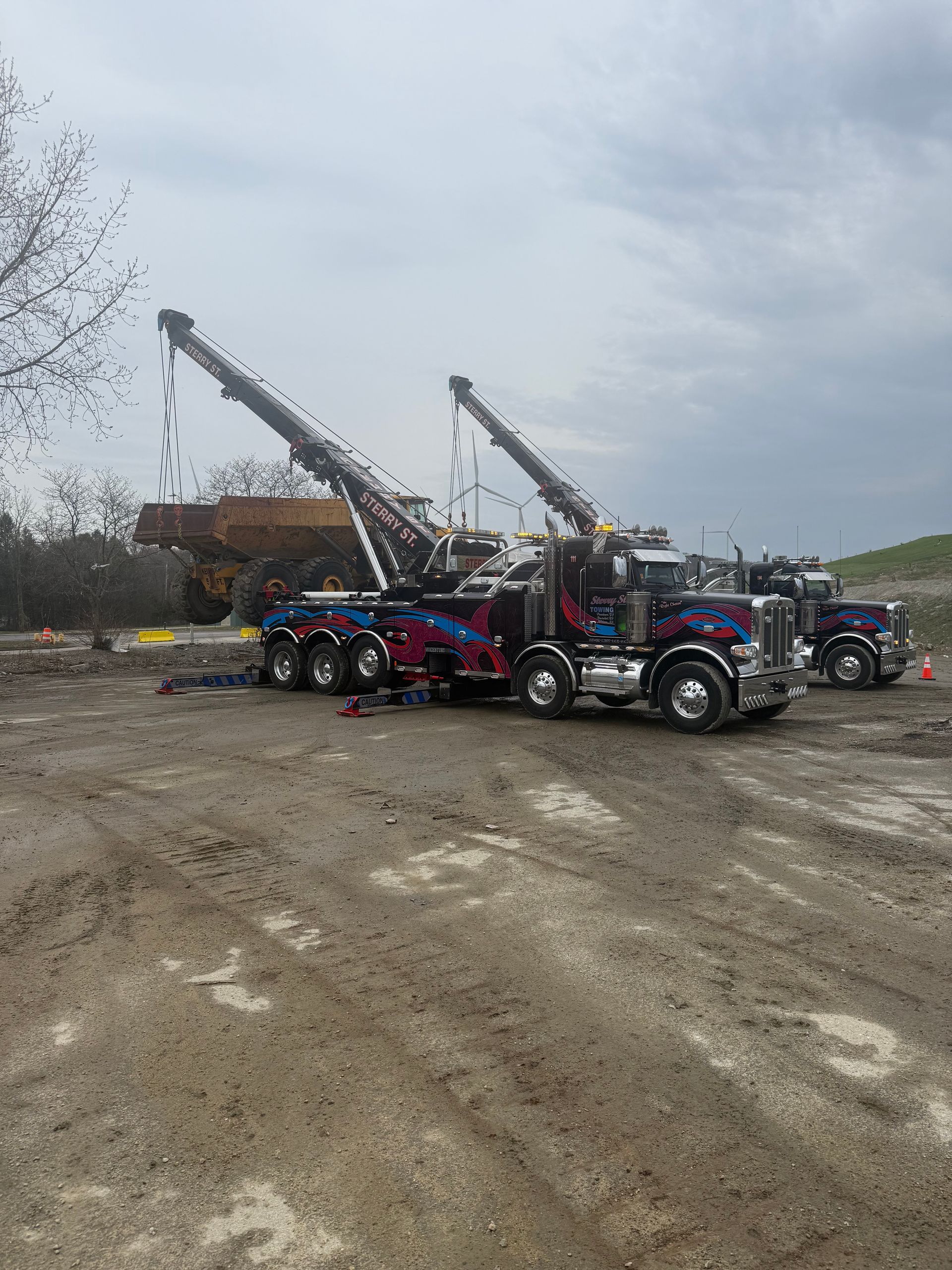 Two tow trucks on a muddy lot lifting a large vehicle, with cloudy sky overhead.