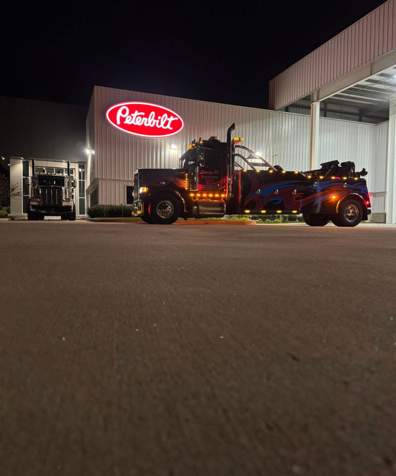 Tow truck and car hauler beside a Peterbilt factory at night, red logo glowing on the building.