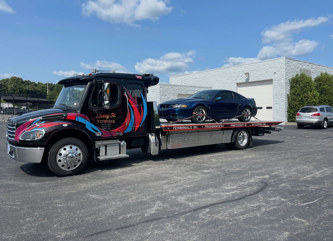 Car hauler truck carrying a black sedan in a parking lot under a blue sky.
