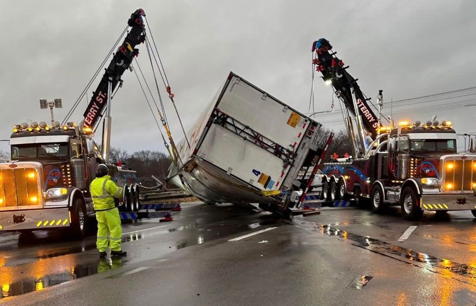 Tow trucks and responders work to upright a toppled semi-trailer on a wet highway.