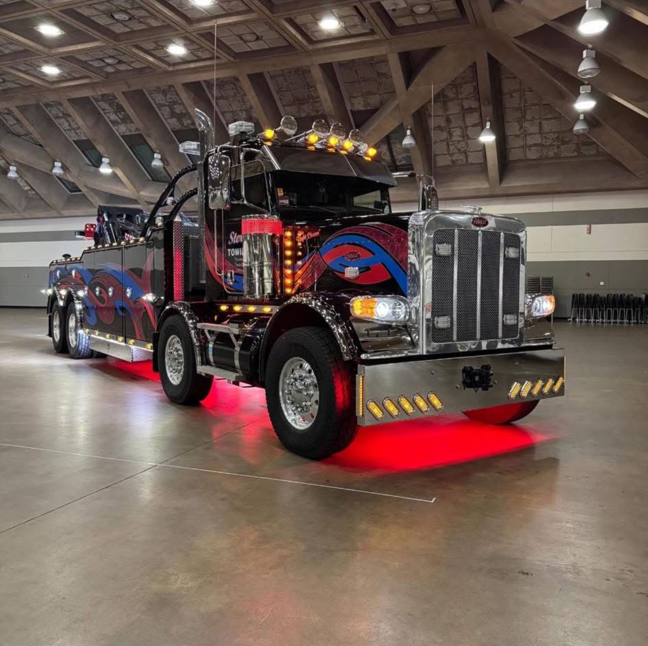 Custom black semi truck with red underglow parked indoors in a large hall.