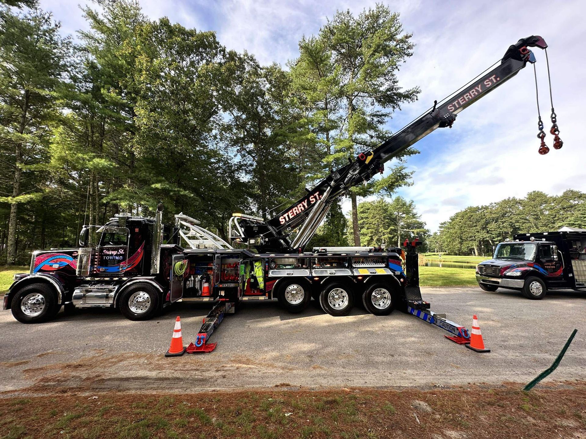 Black crane truck with extended boom on a gravel lot beside trees and parked vehicles.