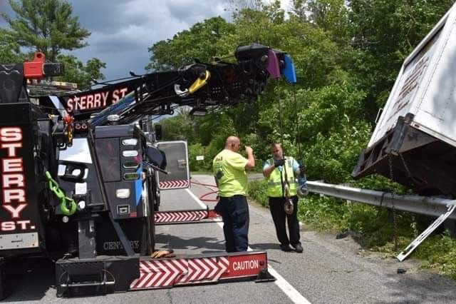 Tow truck crew clearing a fallen roadside sign near a damaged guardrail on a wooded highway.