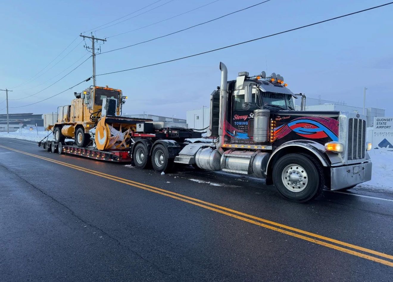 Two large semi trucks hauling heavy equipment on a wet road in a snowy rural area.