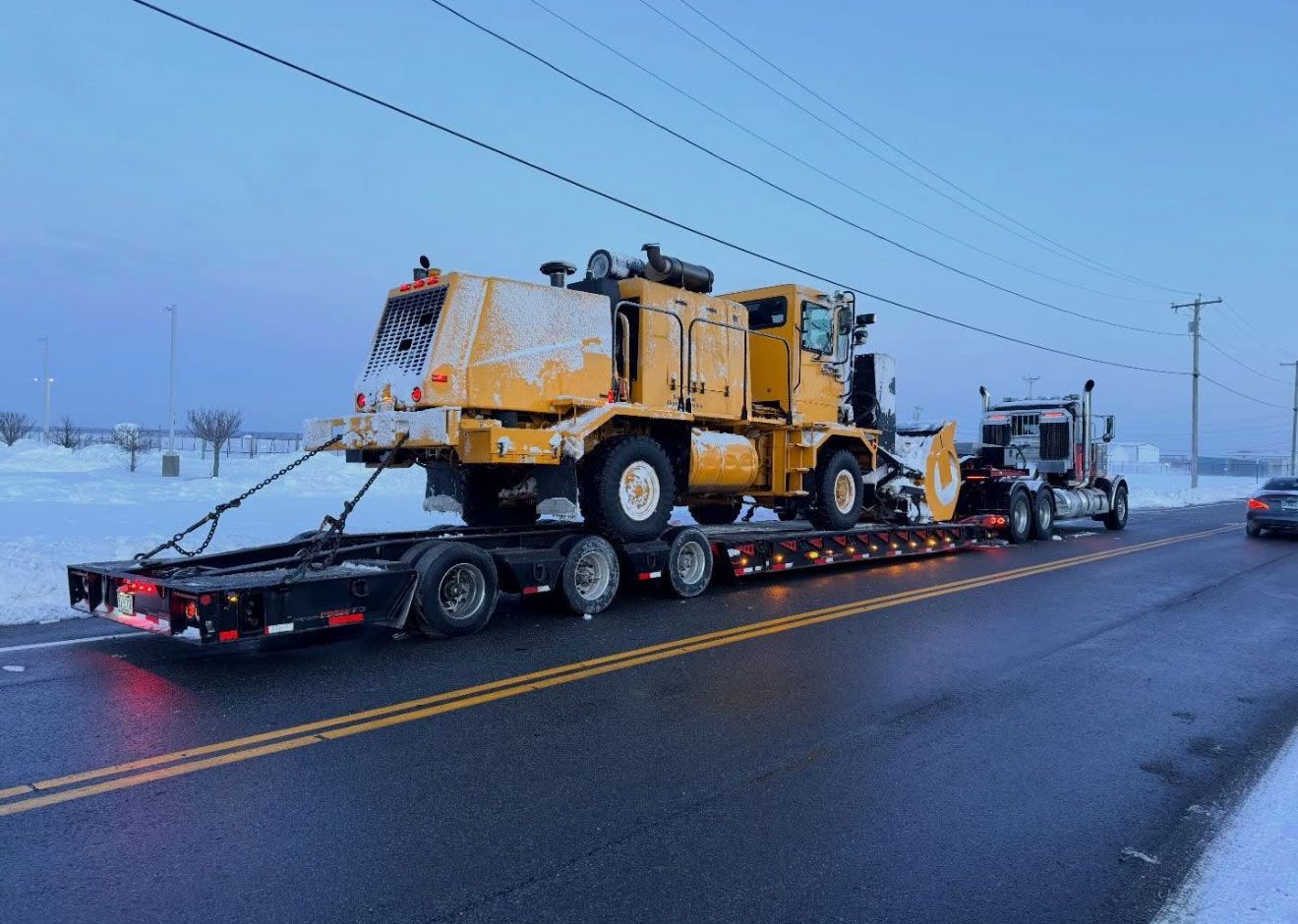 Heavy yellow construction truck on a flatbed trailer being hauled down a wet road at dusk.