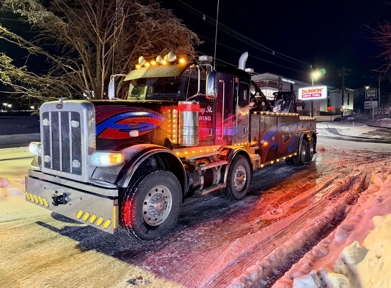 Large red tow truck parked on a snowy roadside at night, lights glowing in the dark.