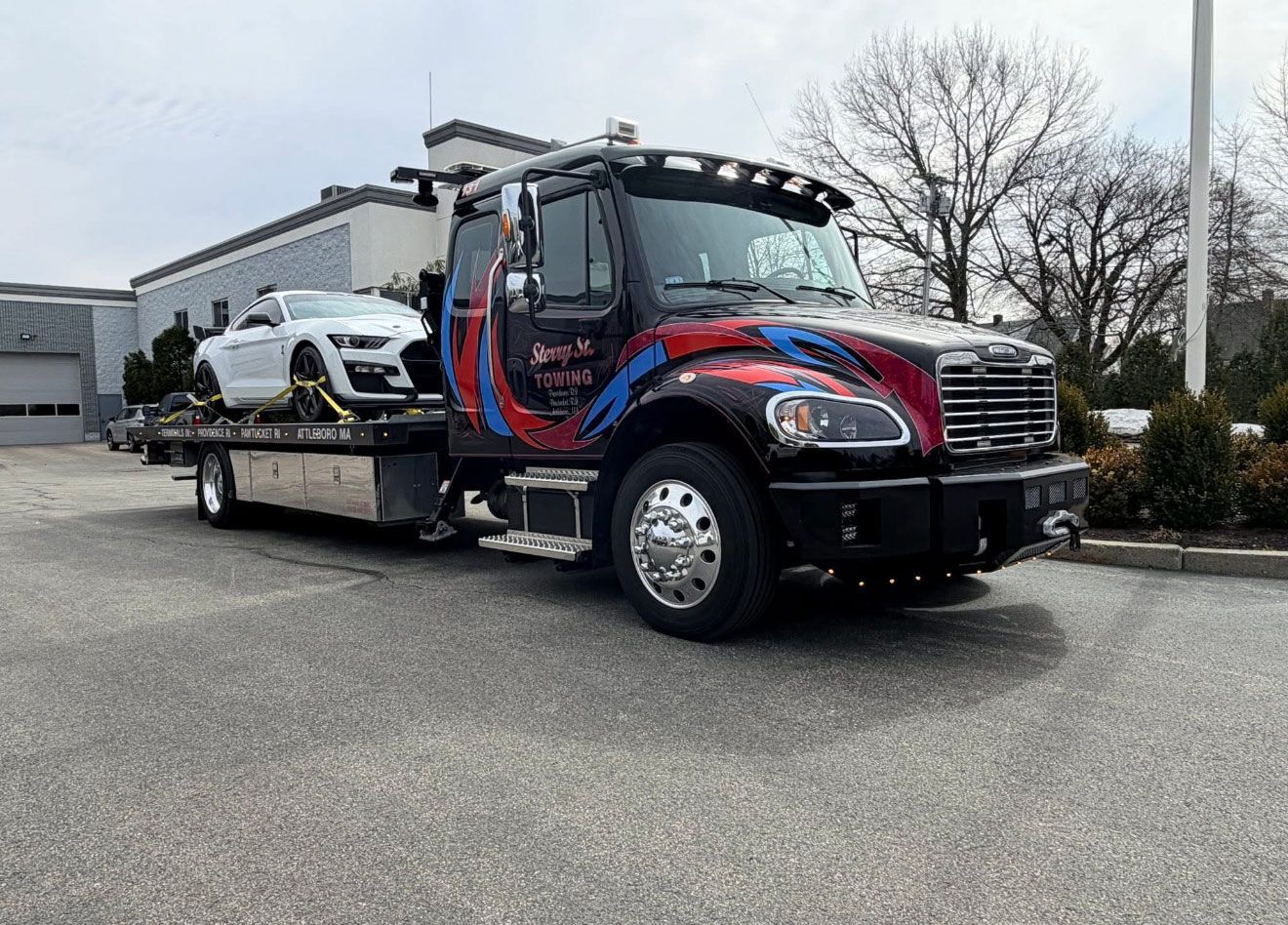 Tow truck carrying a white car on a flatbed in an outdoor parking lot.