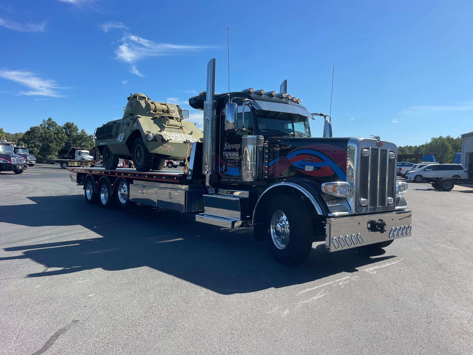 Black semi-truck hauling a military vehicle on a flatbed trailer in a parking lot.
